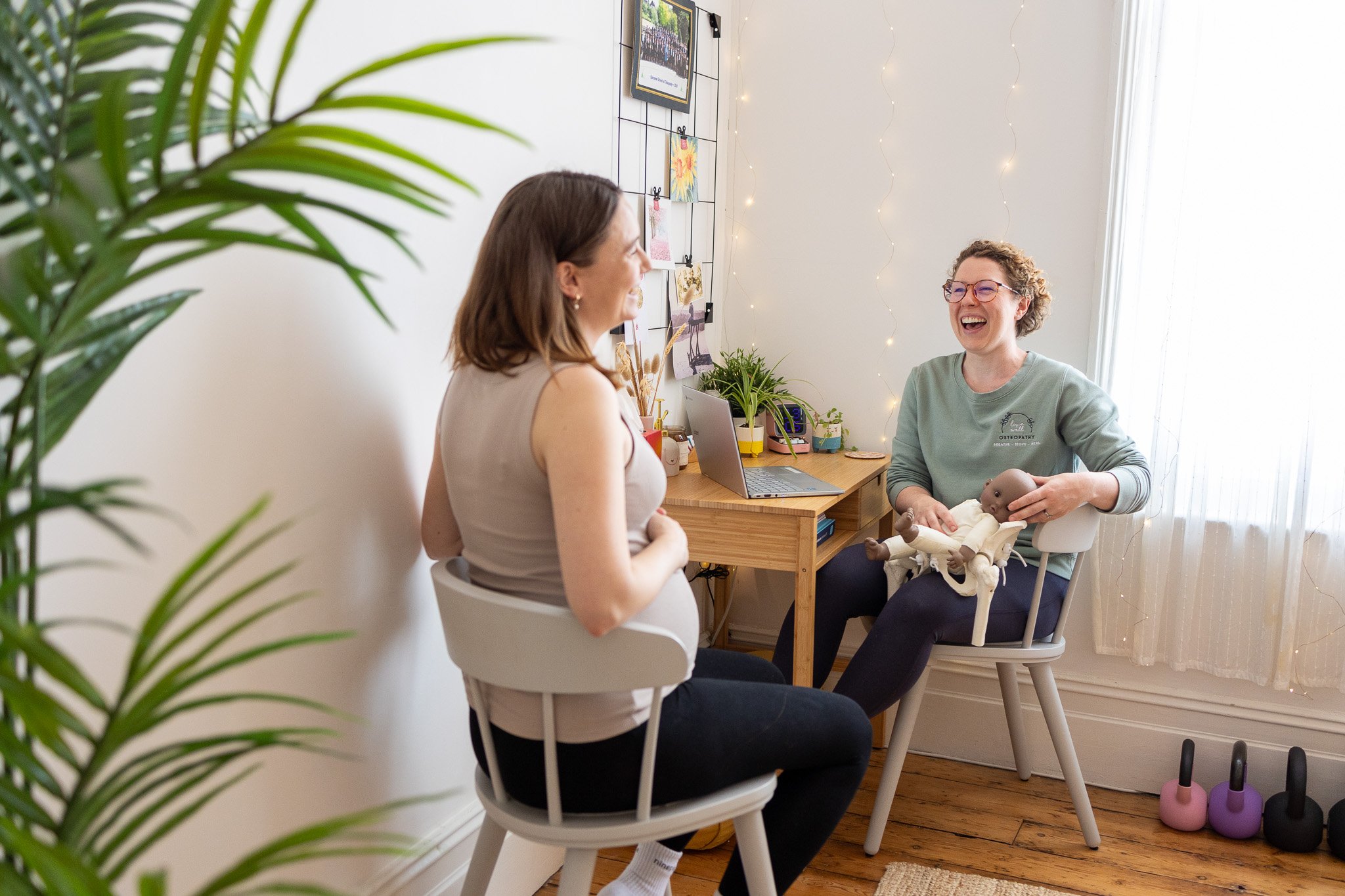A female Osteopath and her pregnant patient sitting and talking in a bright room. The Osteopath is holding a model of a pelvis. Both woman are laughing. There is a small desk with a laptop and plants in the background.