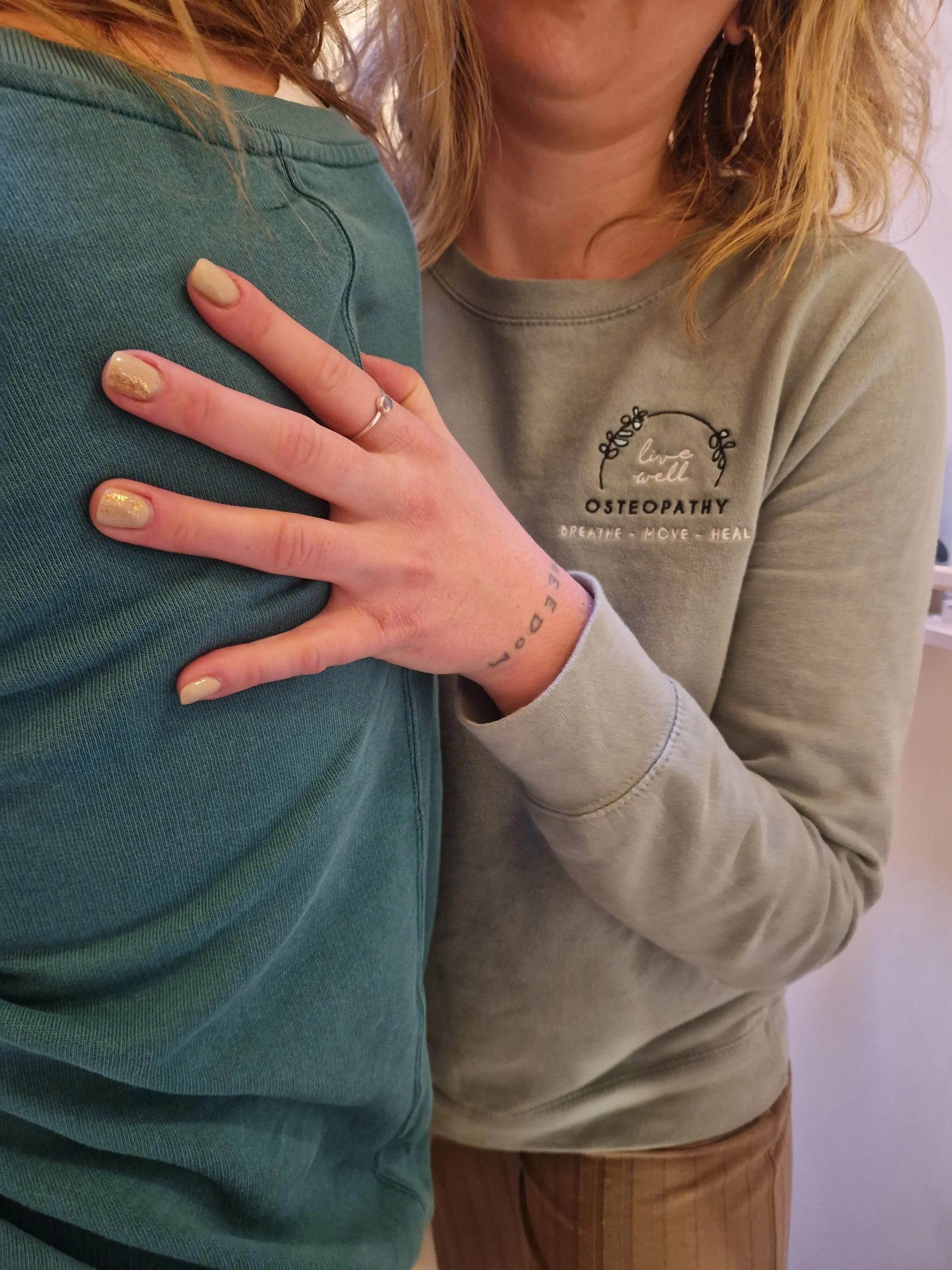 A female Osteopath with blonde hair wearing a grey jumper, performing a treatment on a patient in a green t-shirt. The Osteopath's hand rests on the patient's back.