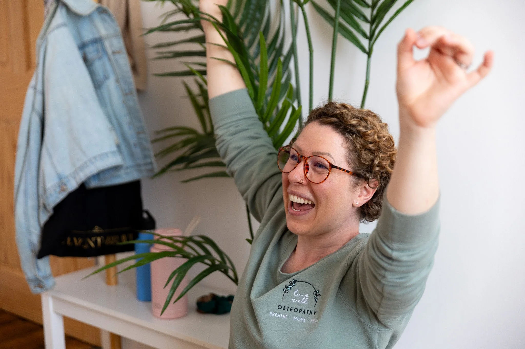 A female Osteopath with curly hair and glasses smiling and stretching her arms above her head in a bright room with plants, wearing a Live Well Osteopathy branded jumper.
