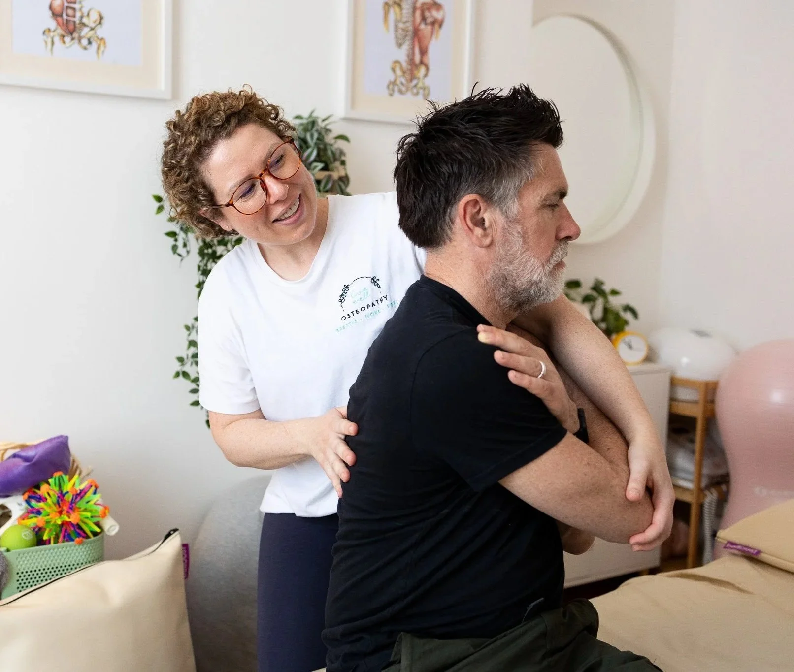 A woman provides osteopathy treatment to a seated man, gently massaging his shoulder and neck in a clinical setting with framed art and plants on the wall.