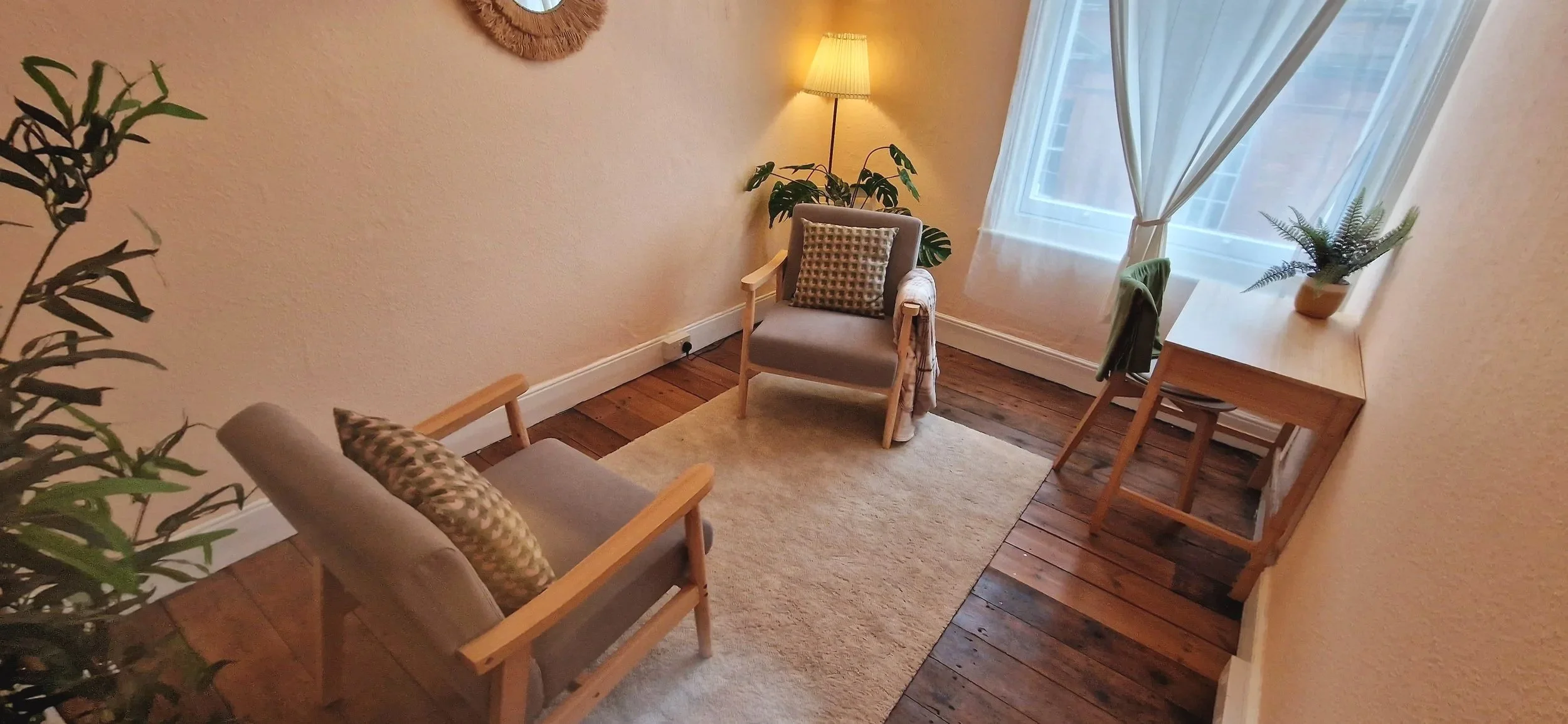A cosy therapy room with two comfy armchairs facing each other, a soft rug, a wooden desk with a plant, and a window with curtains. A lamp provides warm lighting.