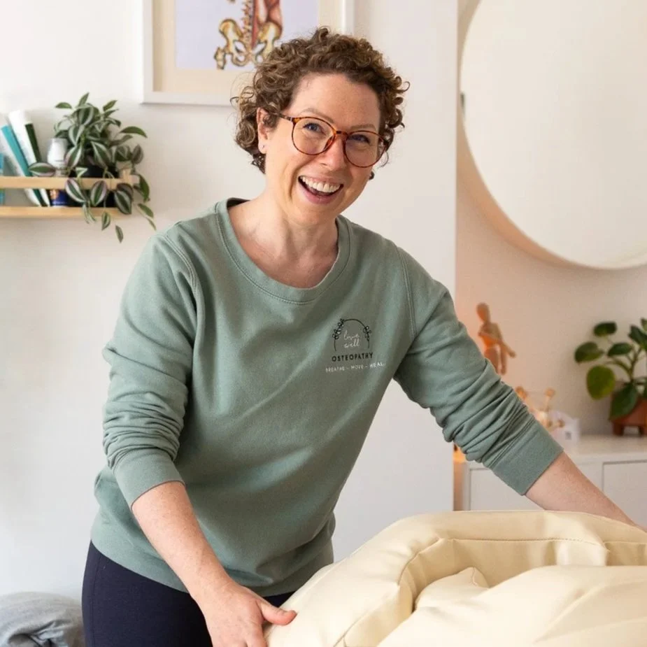 A female Osteopath with curly hair and glasses smiling and leaning over a treatment couch in a brightly lit room with plants, artwork, and soft décor.