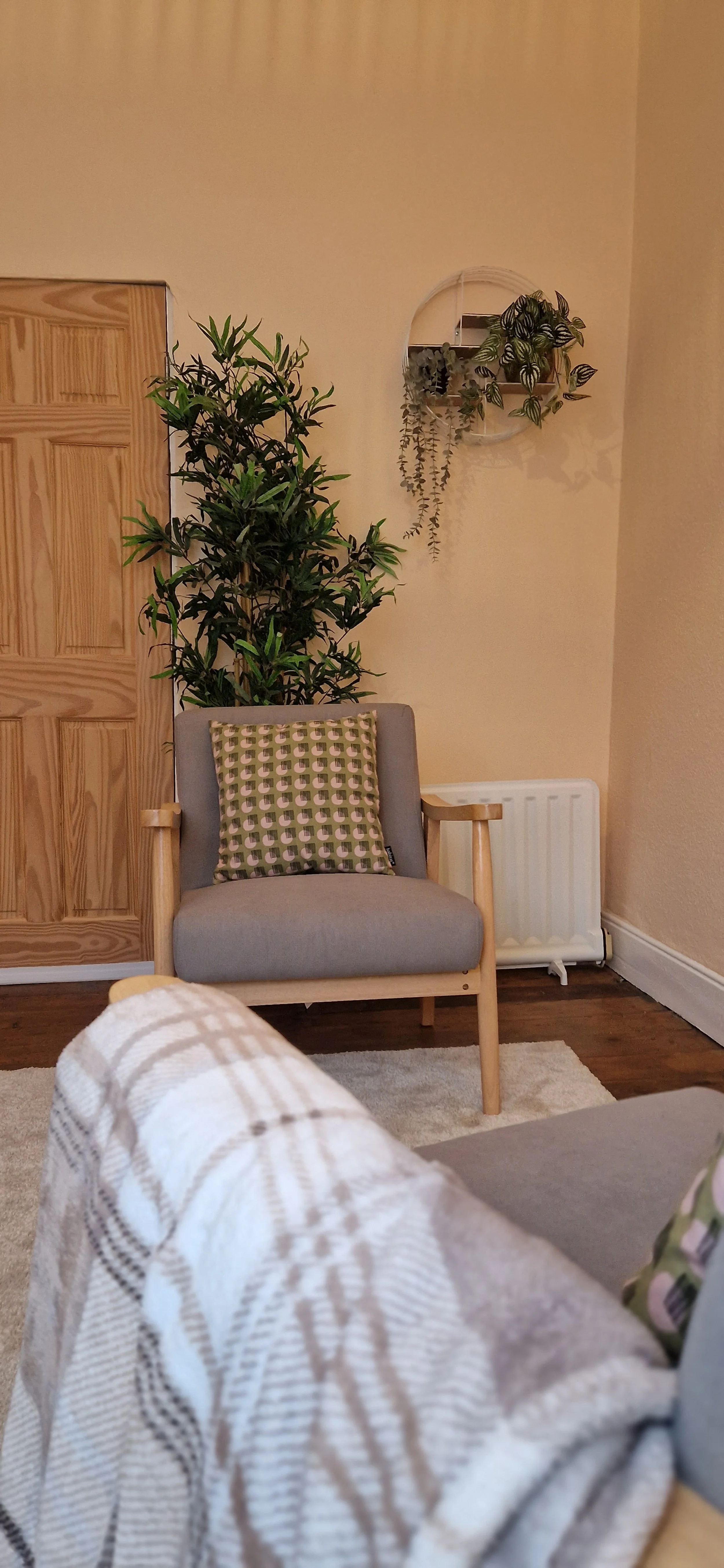 A cosy therapy room with two grey armchairs, a patterned blanket, a large green plant behind one of the chairs, a white wall with a decorative mirror and hanging plants, a wooden door, and a radiator.