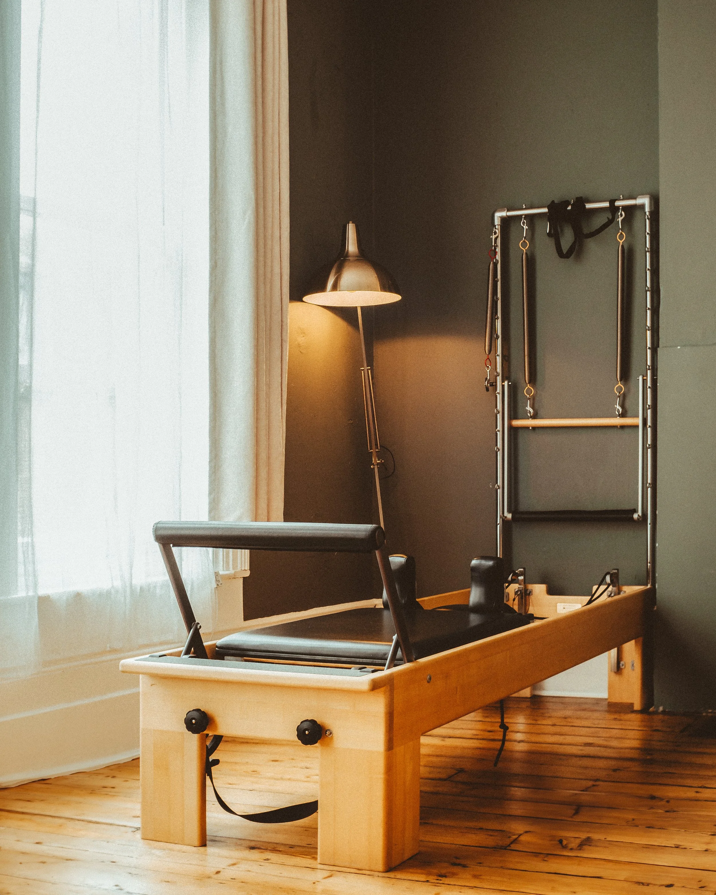 A Pilates reformer machine with a wood frame, black padding, and adjustable foot bar, situated near a large window with white curtains in a room with dark green walls, a floor lamp, and a wall-mounted exercise frame.