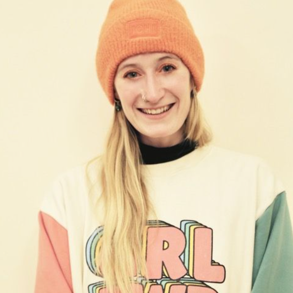 A young woman with long blonde hair, wearing an orange beanie, white jumper with colourful text, and a nose piercing, smiling in front of a plain light background.
