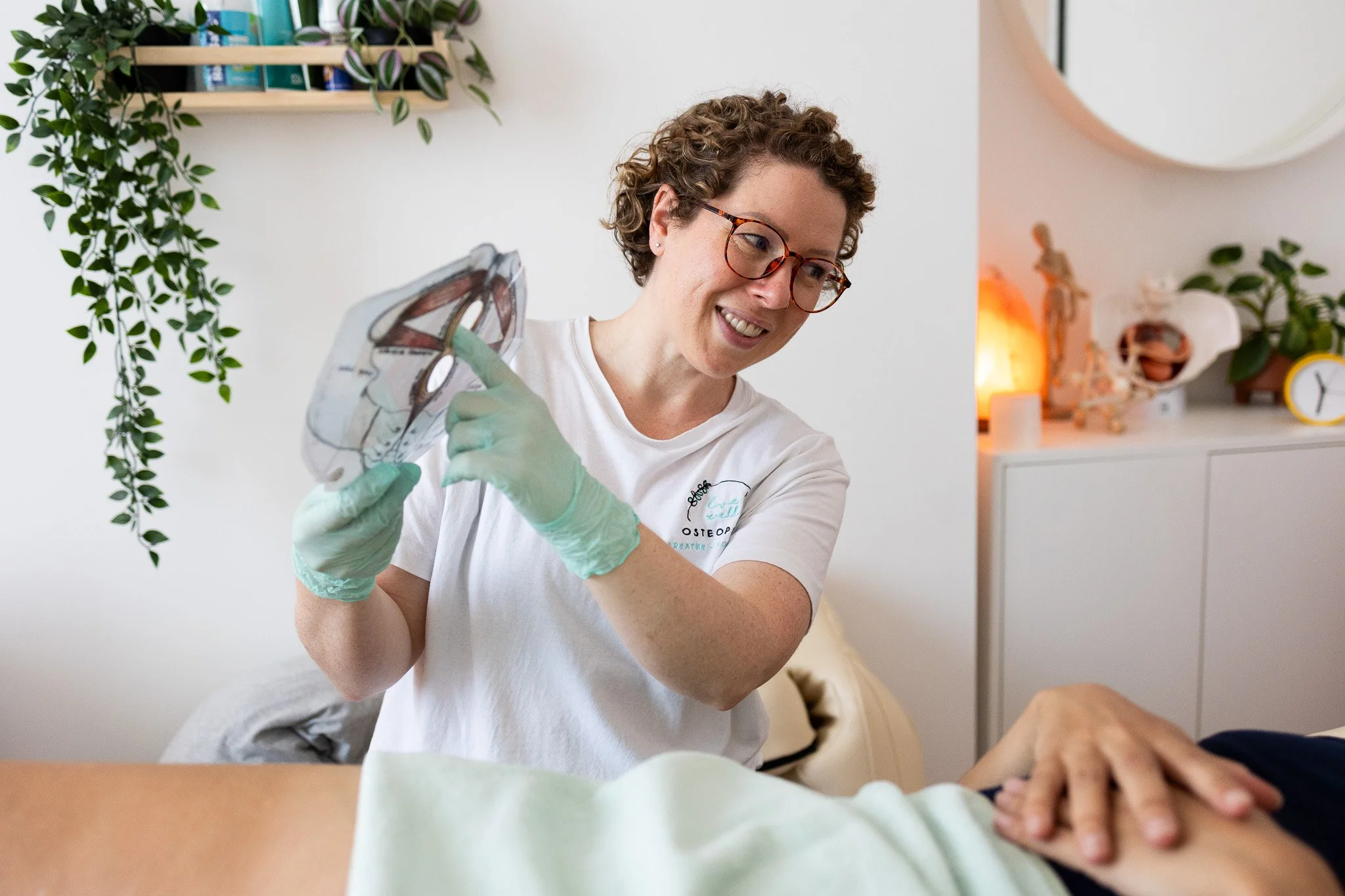 A female Osteopath in medical gloves holding an anatomical model, smiling at a patient lying on a treatment couch.