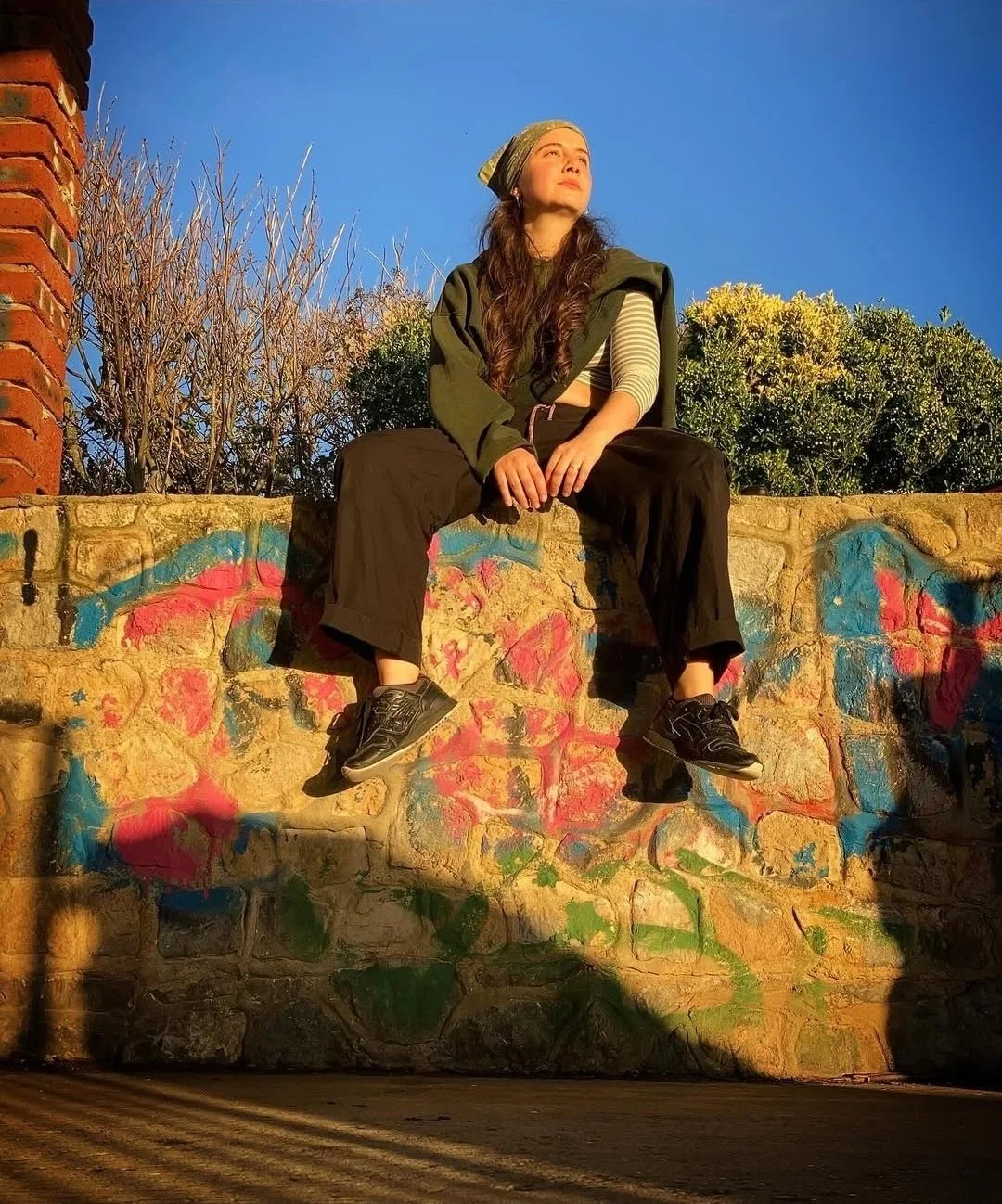 A woman with long hair sitting on a graffiti-covered stone wall at sunset, looking upward with a determined expression, wearing black trousers, a green jacket, sneakers, and a striped long-sleeve shirt, with trees and a blue sky in the background.