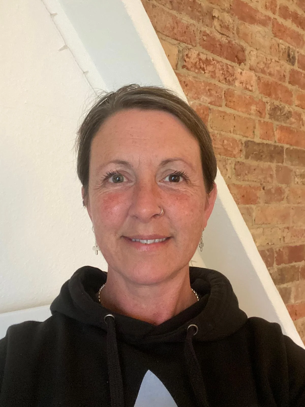 A woman with short brown hair, light skin, and a nose piercing, smiling at the camera. She is wearing earrings, a black hoodie, and a necklace. The background shows a white and brick wall.