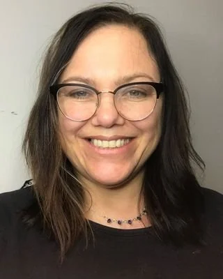 Lyn smiles at the camera. She is wearing glasses and a black top, standing against a white wall.