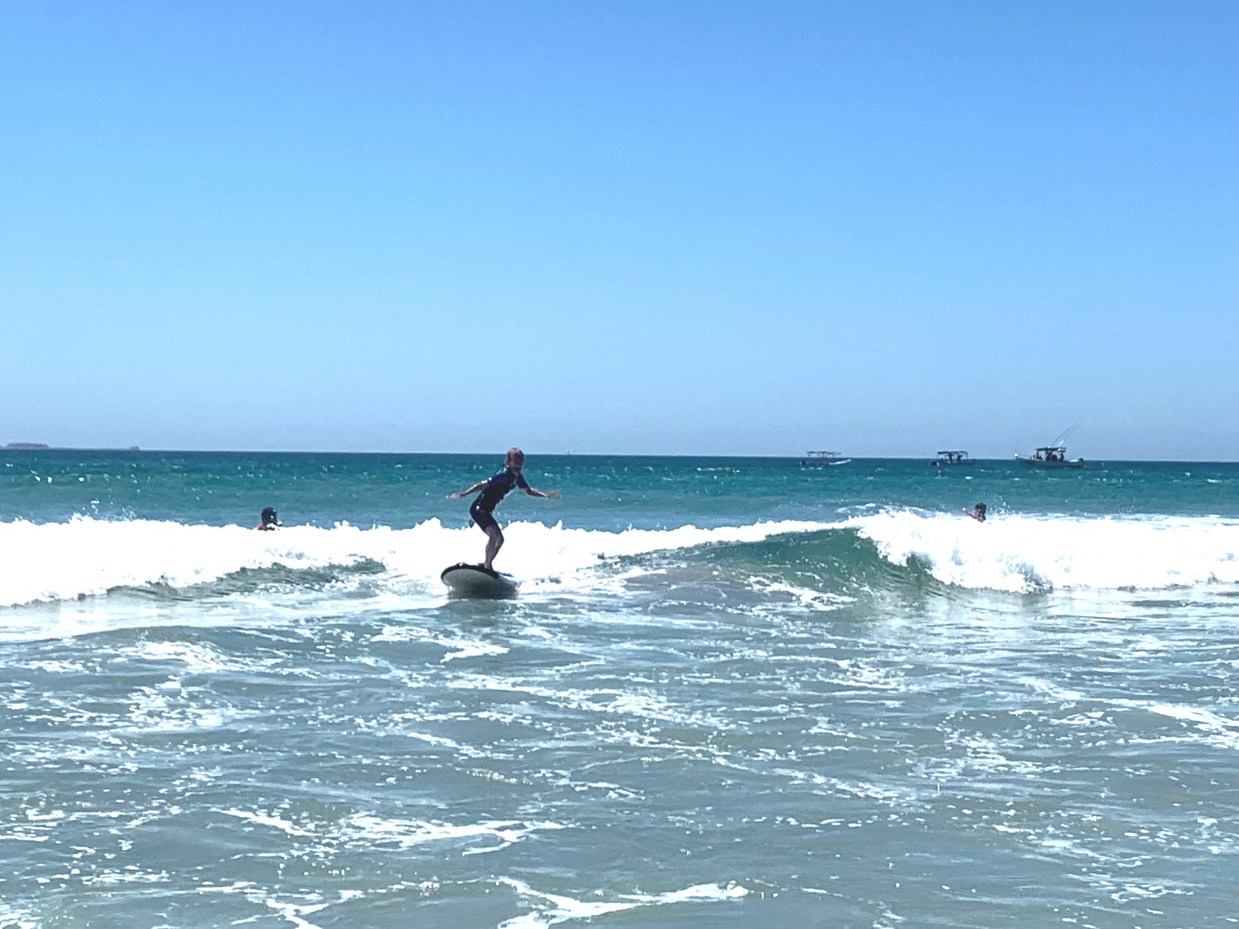 A person riding a surfboard on the ocean waves with others in the water and boats in the background on a sunny day.