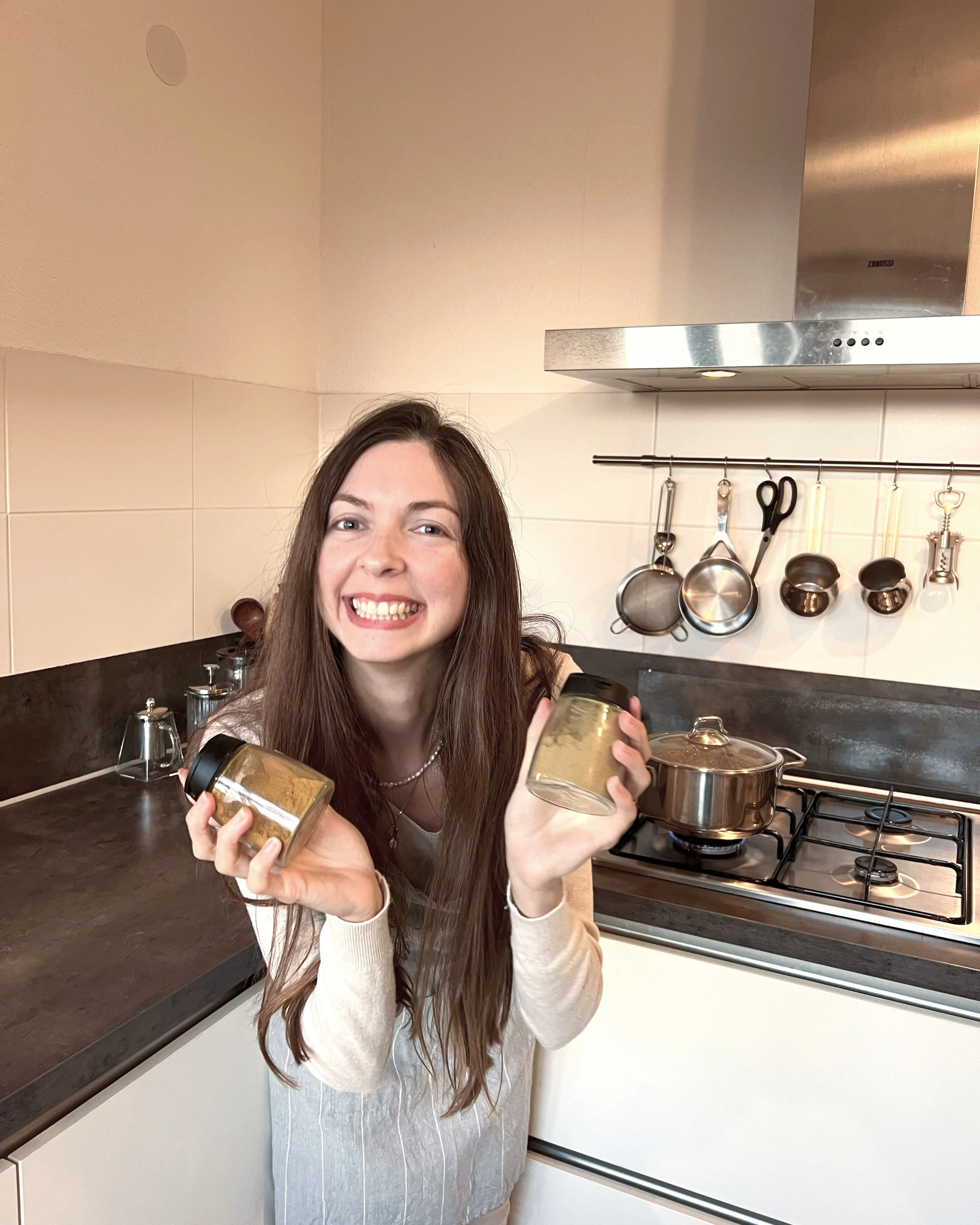 A woman smiling and holding two jars of spice in a kitchen with pots and utensils hanging on a wall behind her.