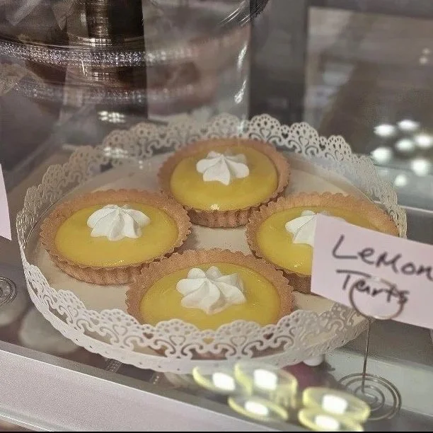 Four lemon tarts topped with whipped cream rosettes on a white lace-style stand in a bakery display case, with a handwritten “Lemon Tarts” label.
