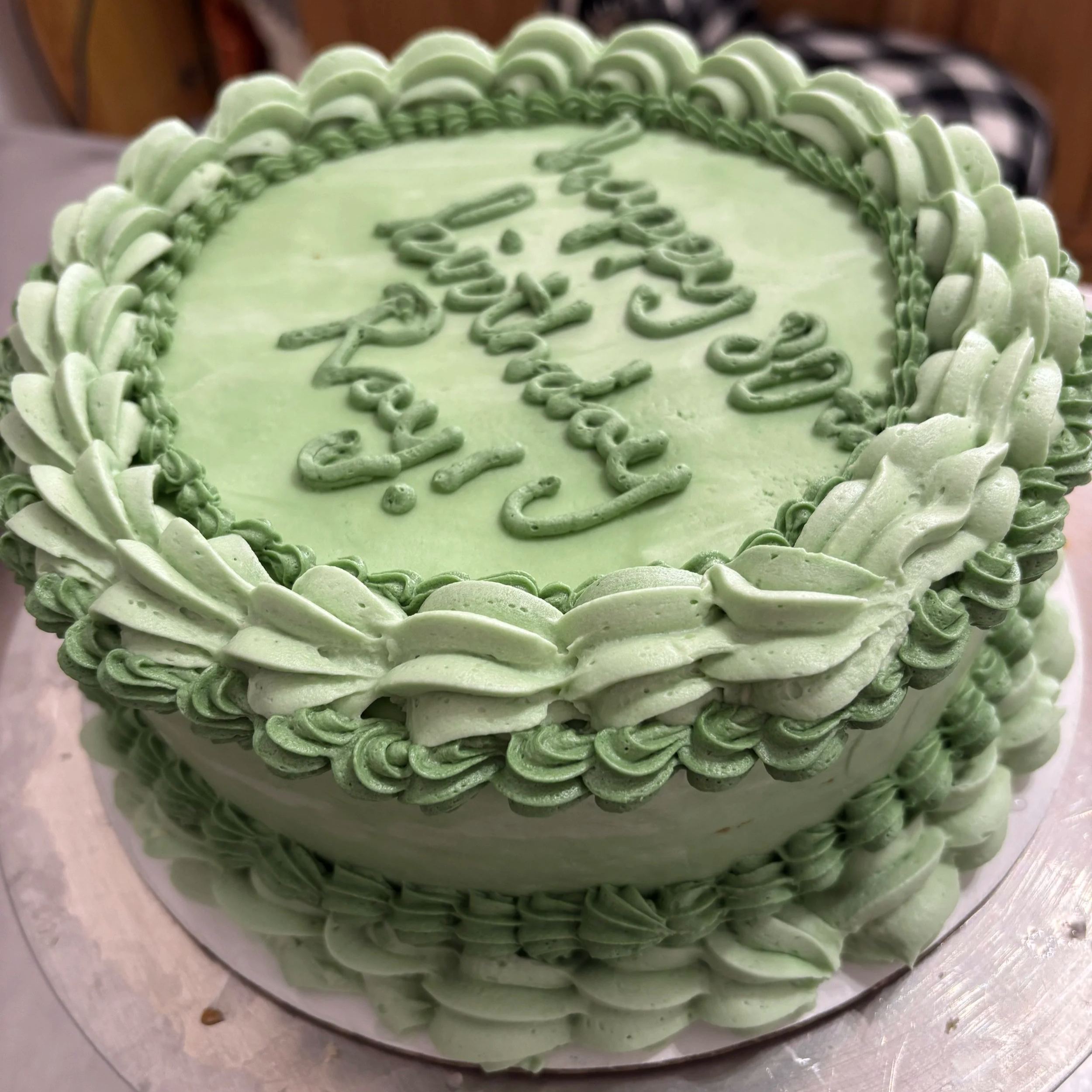 Round birthday cake in two shades of green buttercream with ruffled shell borders and “Happy Birthday” piped in green script on top.