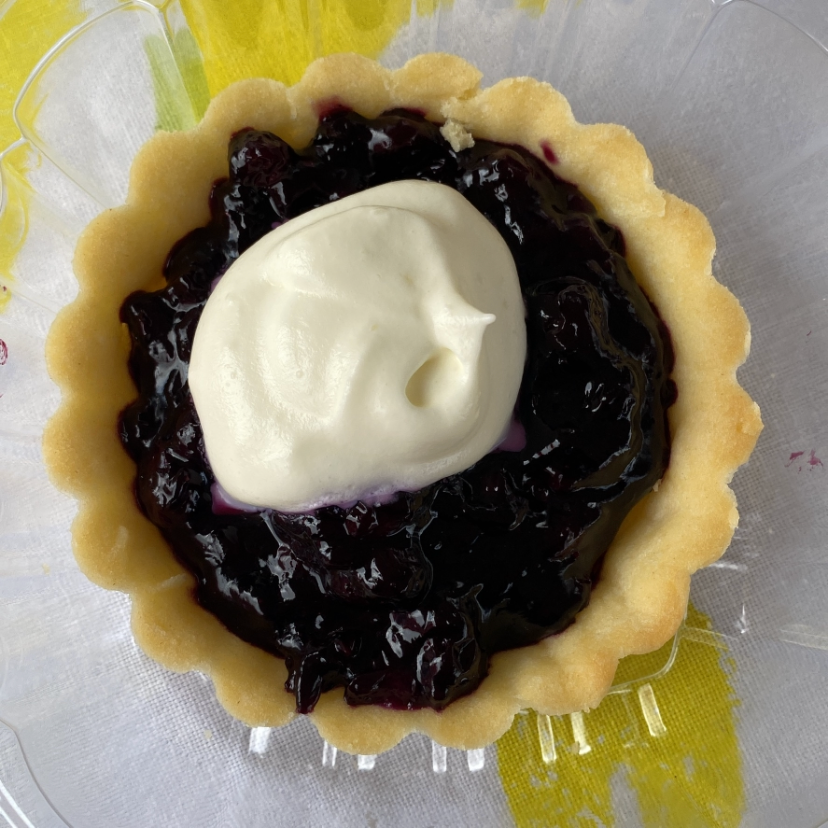 Single tart with a golden fluted crust, dark blueberry filling, and a dollop of whipped cream on top, on a clear plate over a yellow-patterned surface.