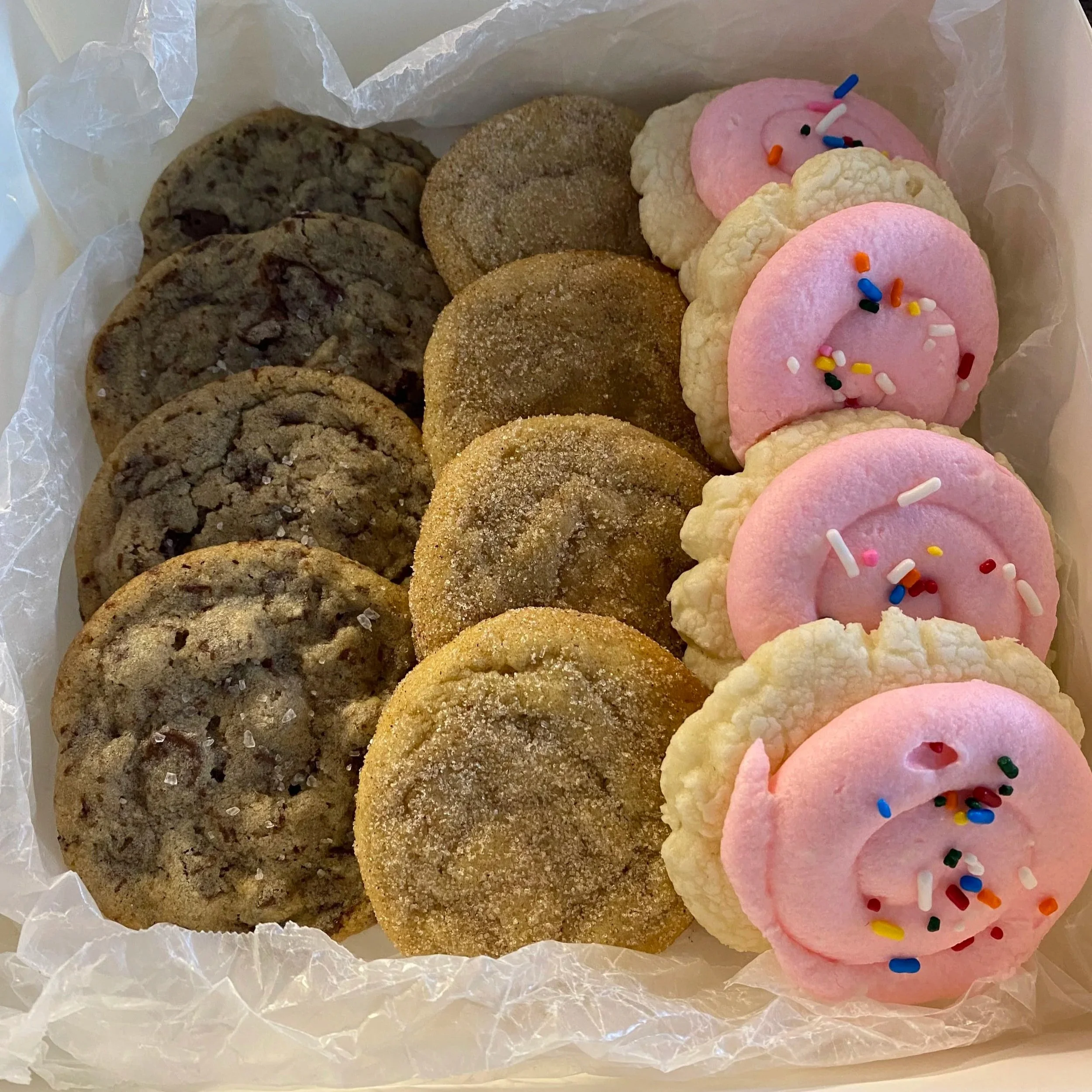 Box of assorted cookies lined with parchment: chocolate chip cookies on one side and round sugar cookies with pink frosting and rainbow sprinkles on the other.