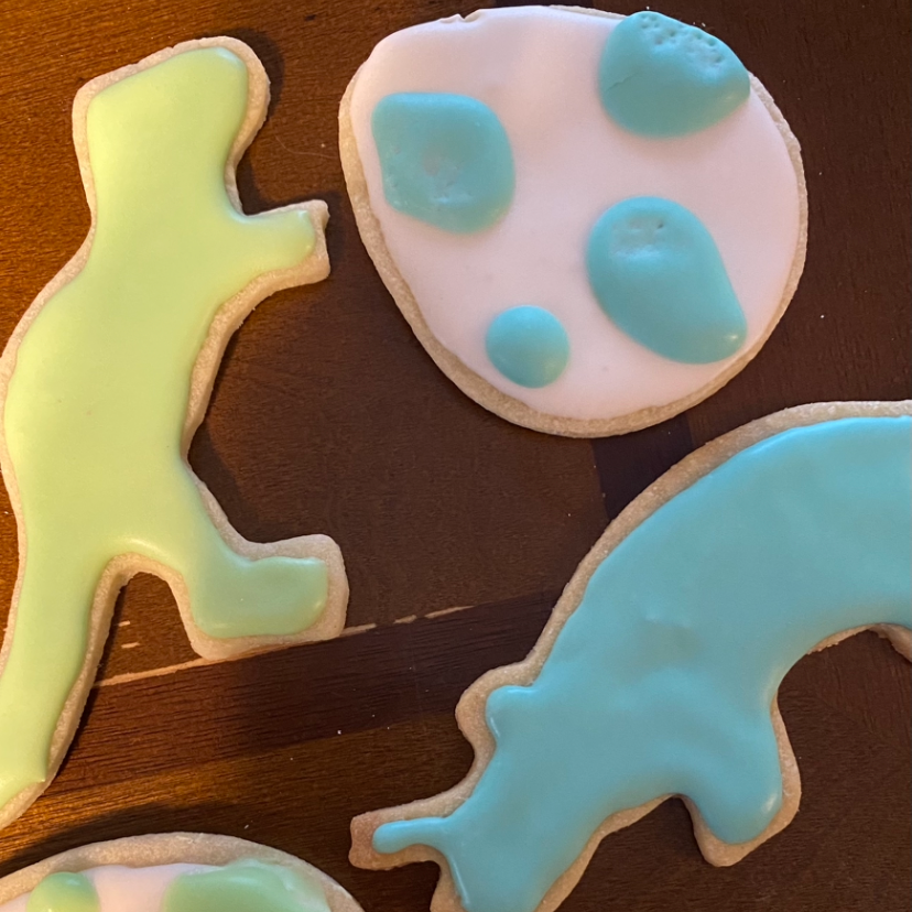 Iced sugar cookies shaped like dinosaurs in pastel yellow-green and blue, next to a pink egg-shaped cookie with blue spots, on a dark wood surface.