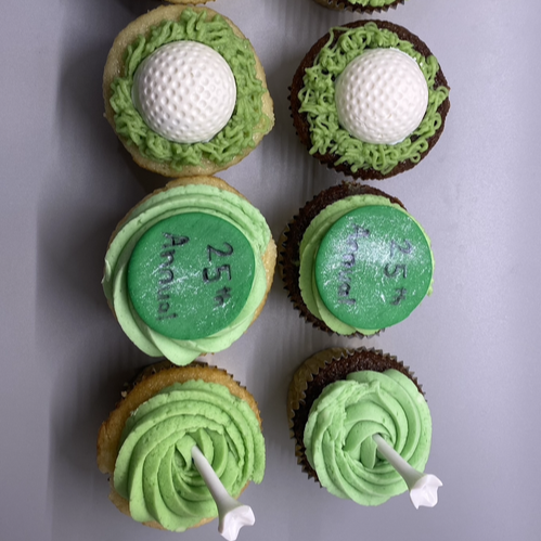 Overhead view of golf-themed cupcakes: some with piped green “grass” frosting and a white golf ball on top, some with green frosting swirls and a golf tee, and some with round green “25” discs.