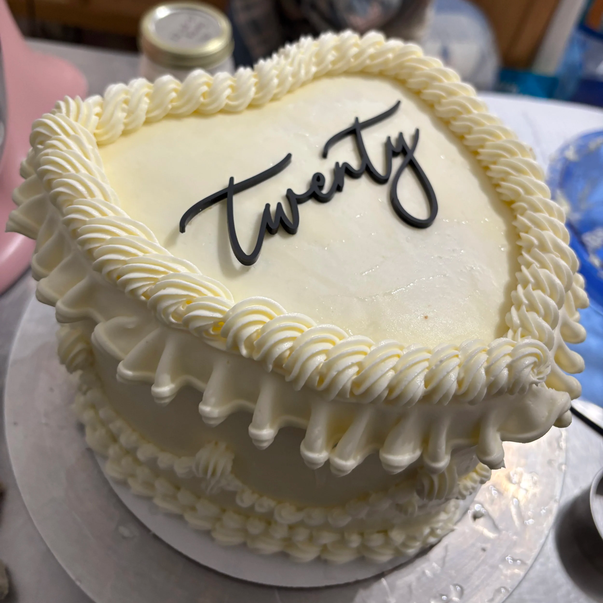Heart-shaped white buttercream cake with vintage piped rope and ruffle borders and a black script “Twenty” cake topper, on a silver cake board.