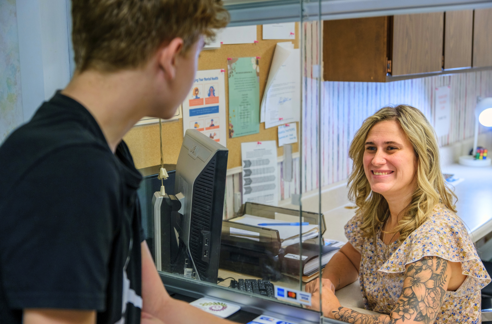 A young adult is looking at a woman smiling sitting at her desk next to a computer.