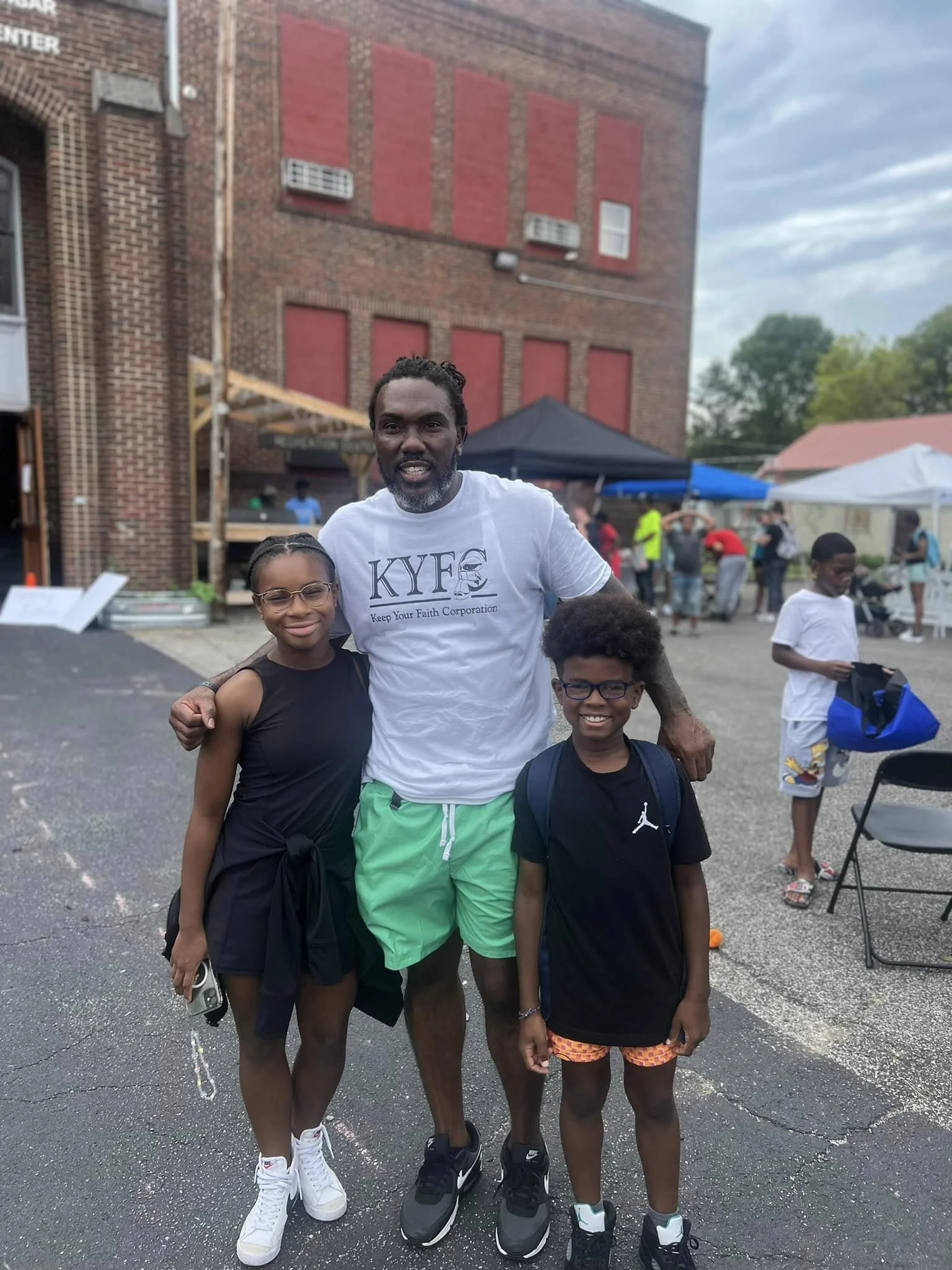 An adult man and two children are standing together outside at a community event.