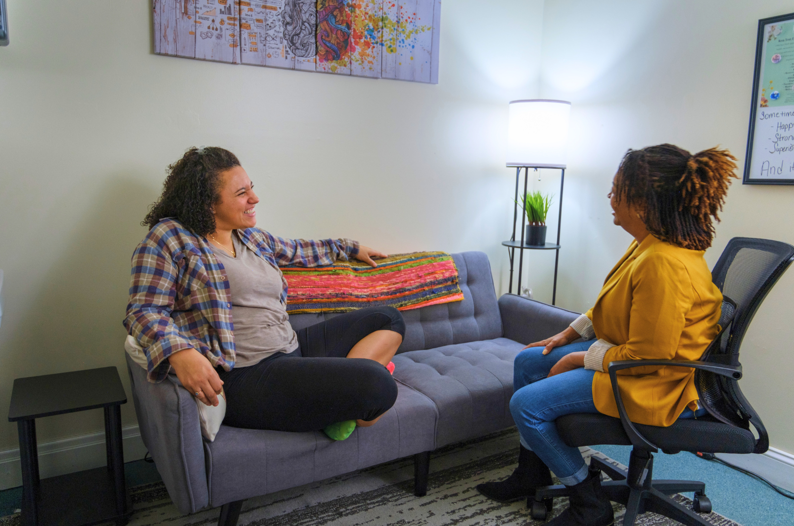 An adult patient is sitting on a couch smiling while looking at their counselor. The counselor is sitting in an office chair.