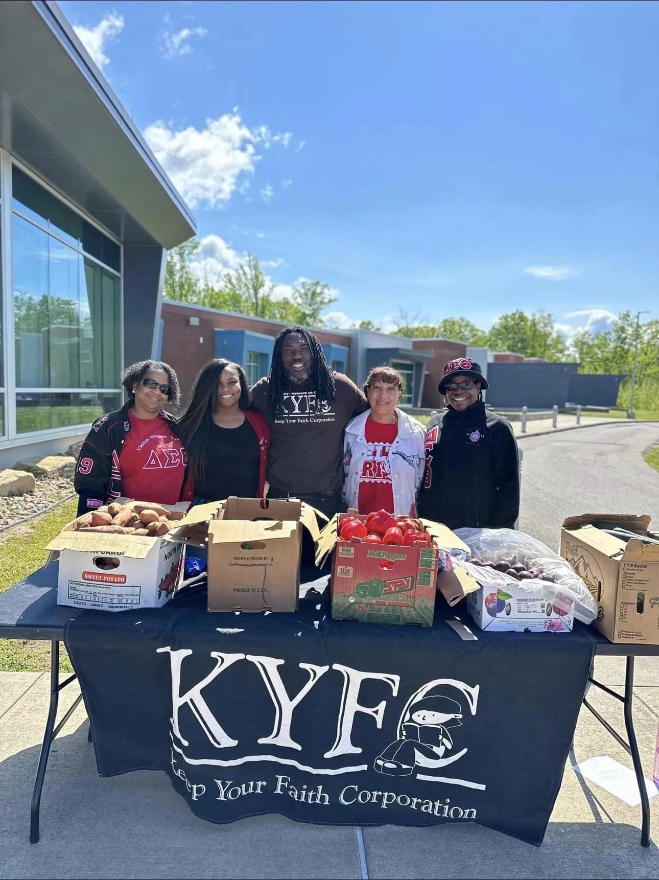 A group of five adults are standing together smiling behind a table that has produce in boxes on top. They are outside on a sidewalk with a building behind them.