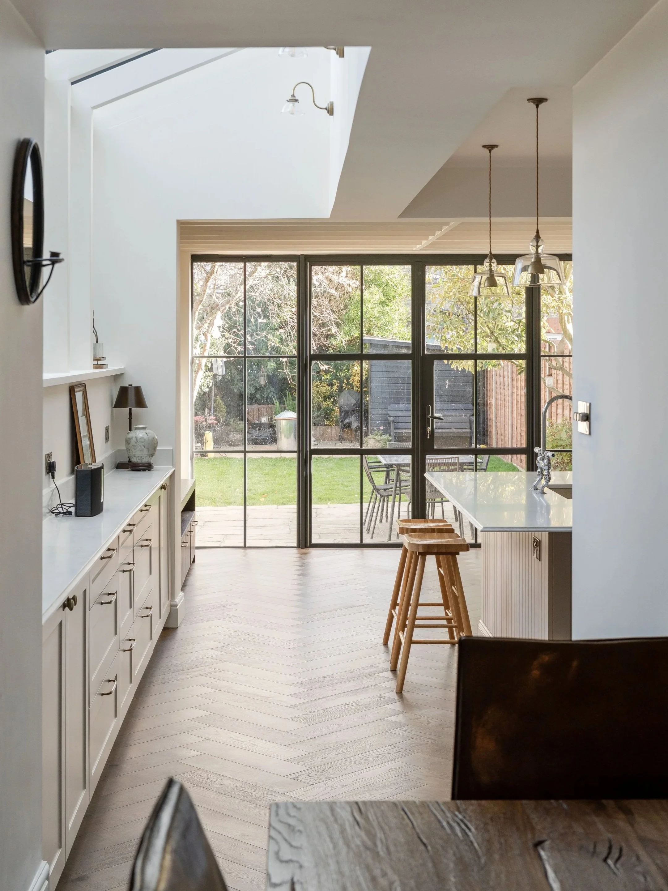 Bright kitchen with white cabinets, a large window and sliding glass doors overlooking a backyard with grass, trees, outdoor furniture, and a shed. Wooden bar stools are at a white kitchen island, and hanging lights are above.