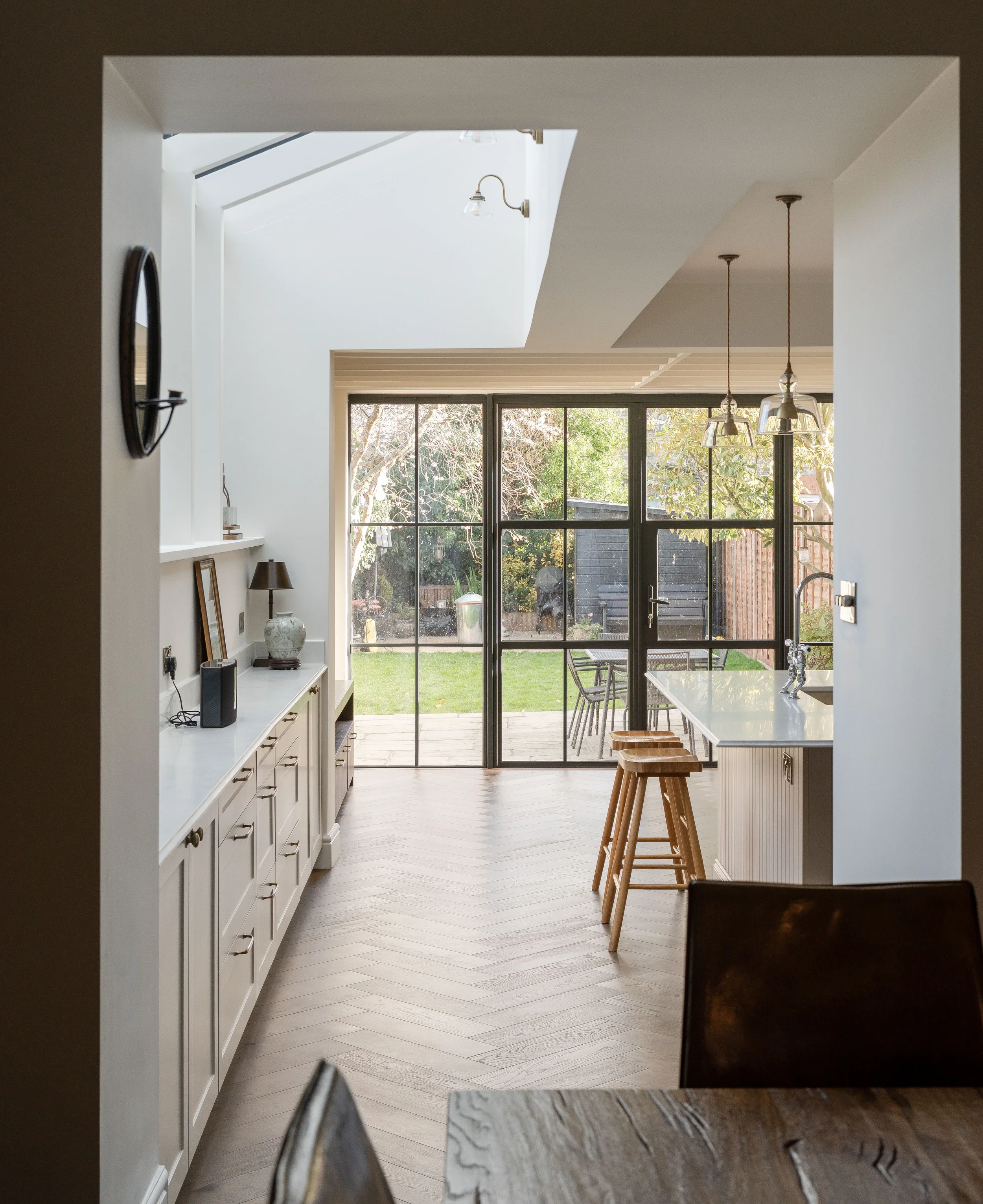 Modern kitchen with white cabinets, wooden bar stools, large glass sliding doors opening to a backyard, and pendant lighting.