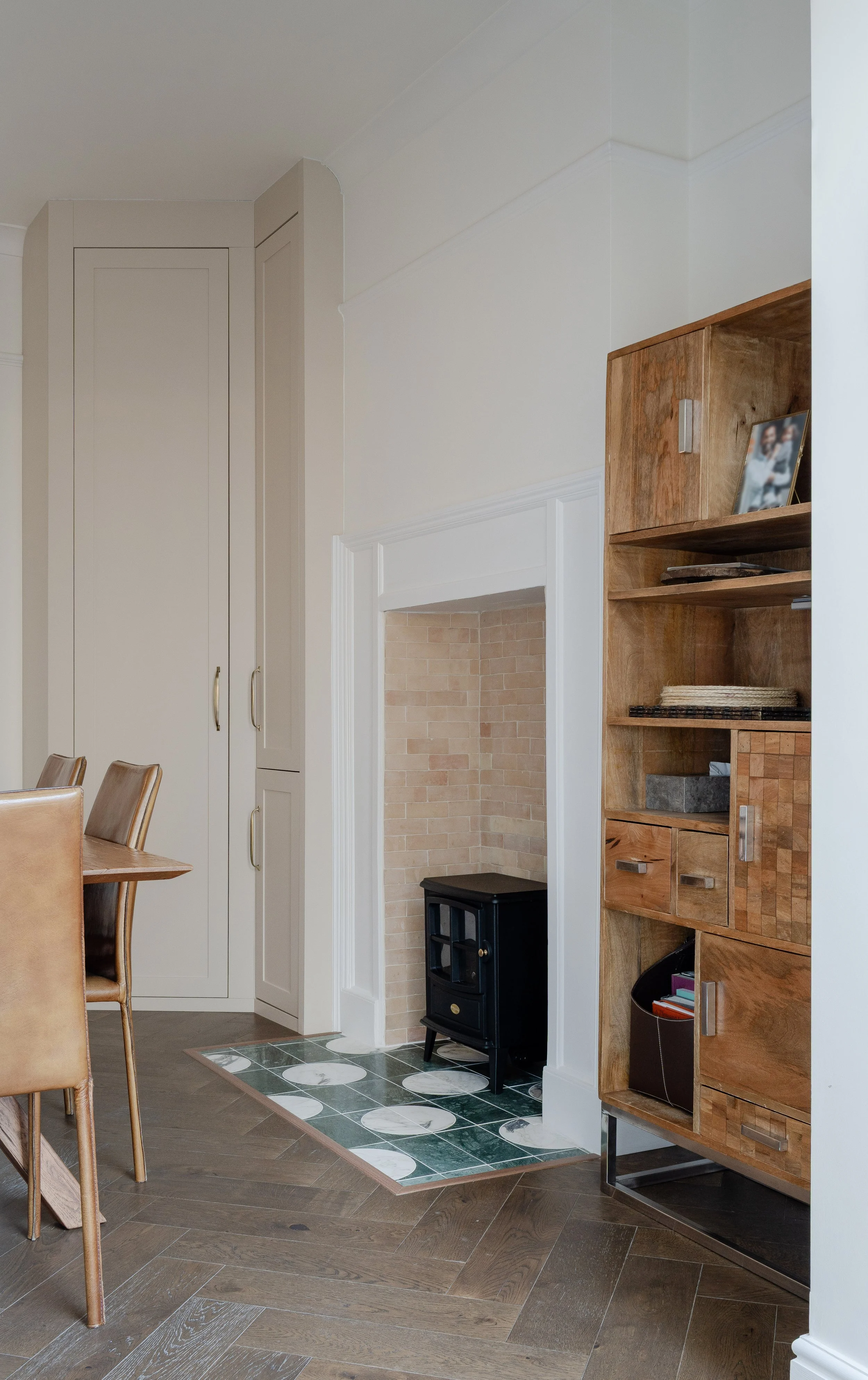 Interior view of a room showing beige cabinetry, a wooden cabinet with books and decor, a small black stove on green and white patterned tile, and a dining table with tan chairs.