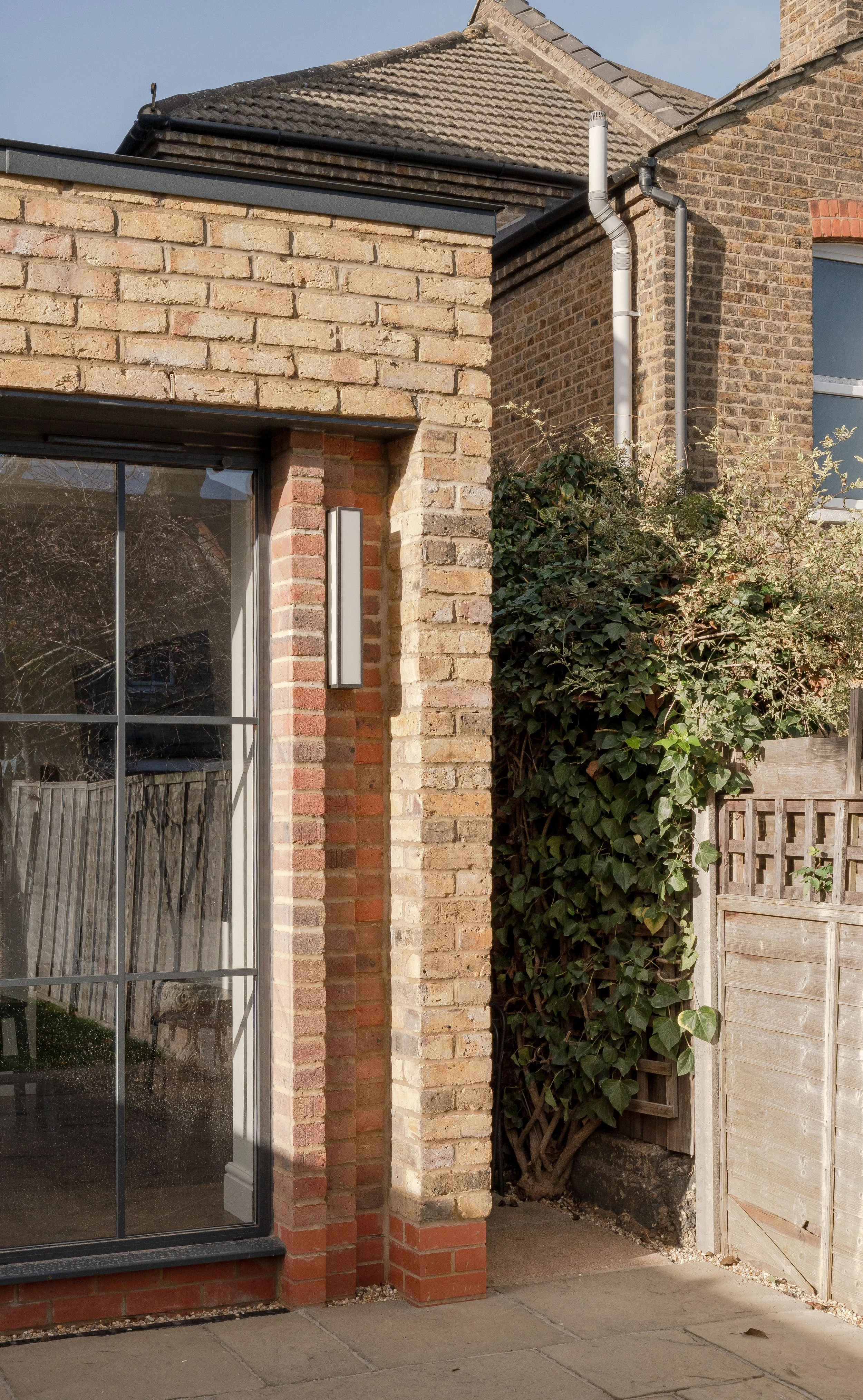Close-up of a brick house corner with a large window, outdoor wall light, and overgrown ivy climbing a wooden fence, with a cloudy sky in the background.