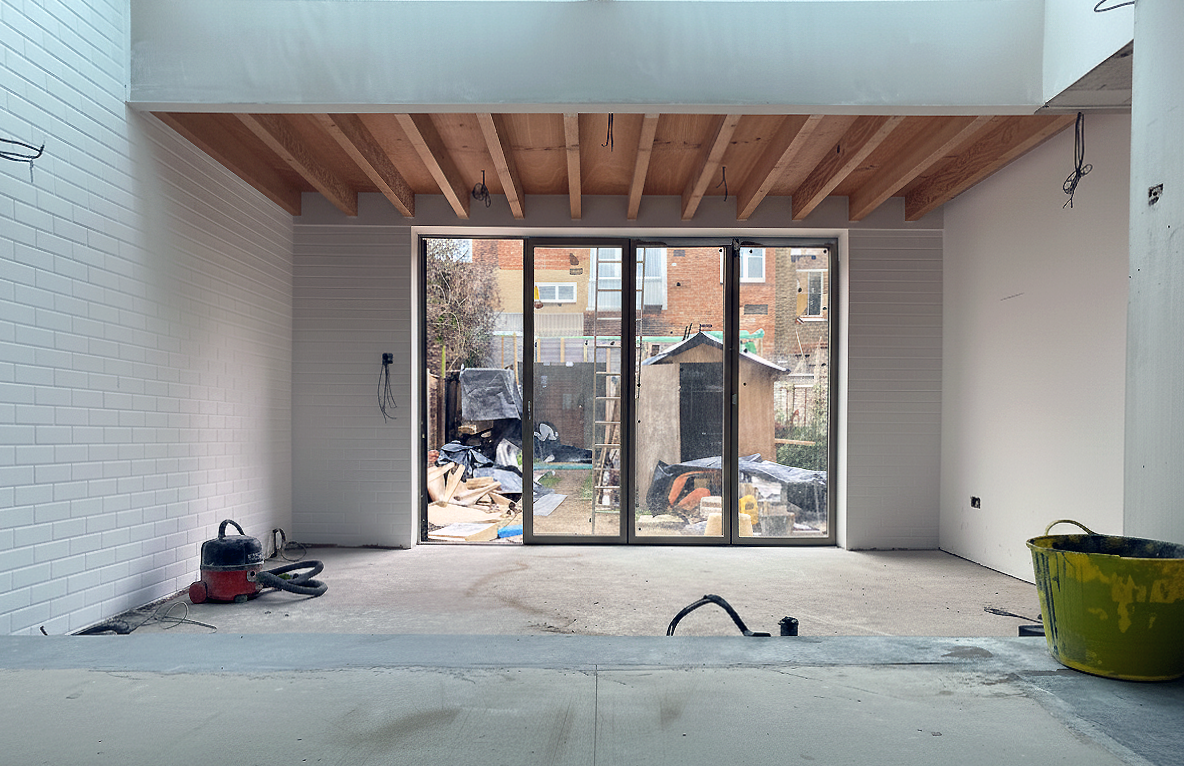 Interior of a room under construction with exposed wooden beams on the ceiling, unfinished flooring, and a large sliding glass door opening to an outdoor area with construction materials and a shed.