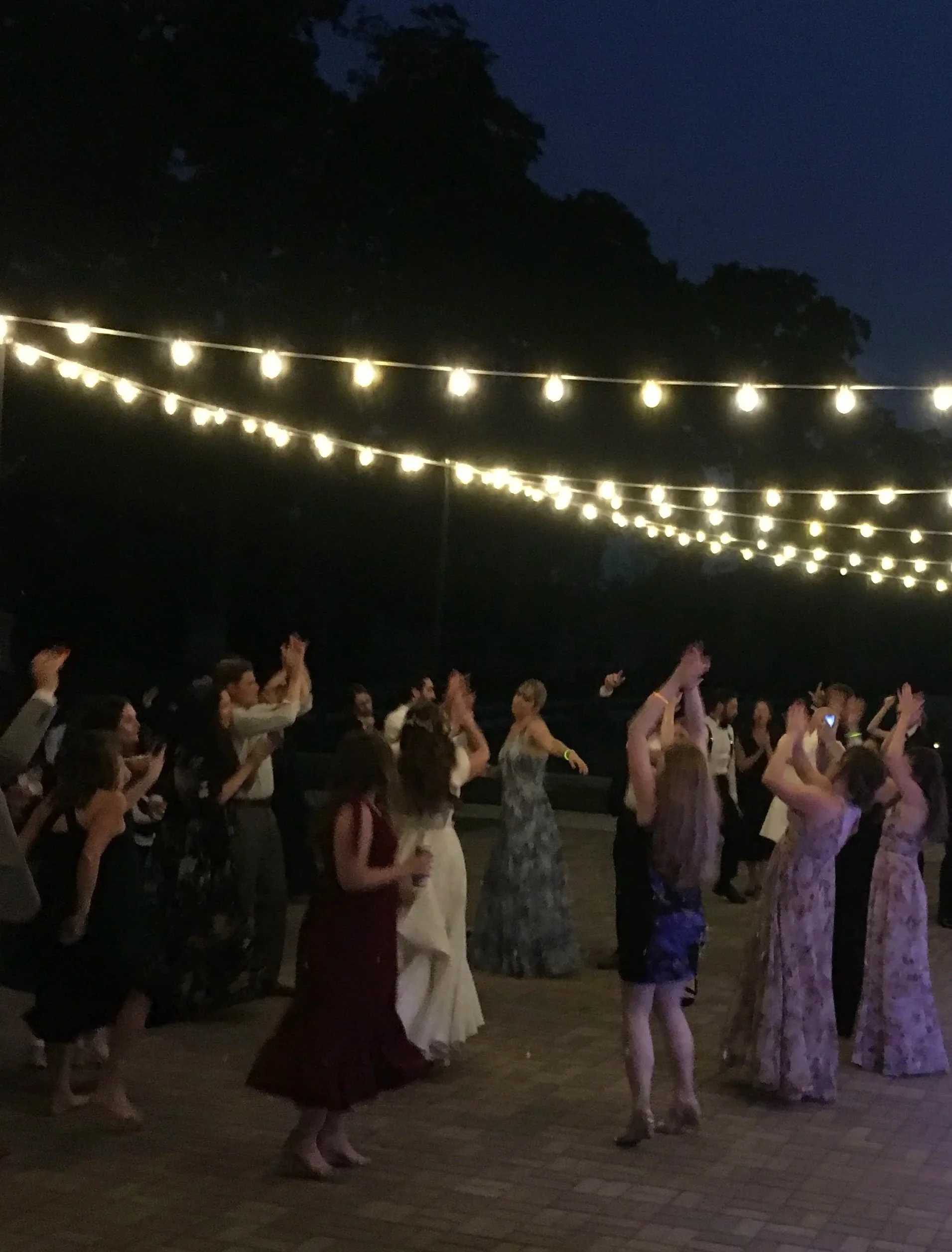 Guests dancing beneath glowing string lights on an estate patio during a summer wedding celebration.