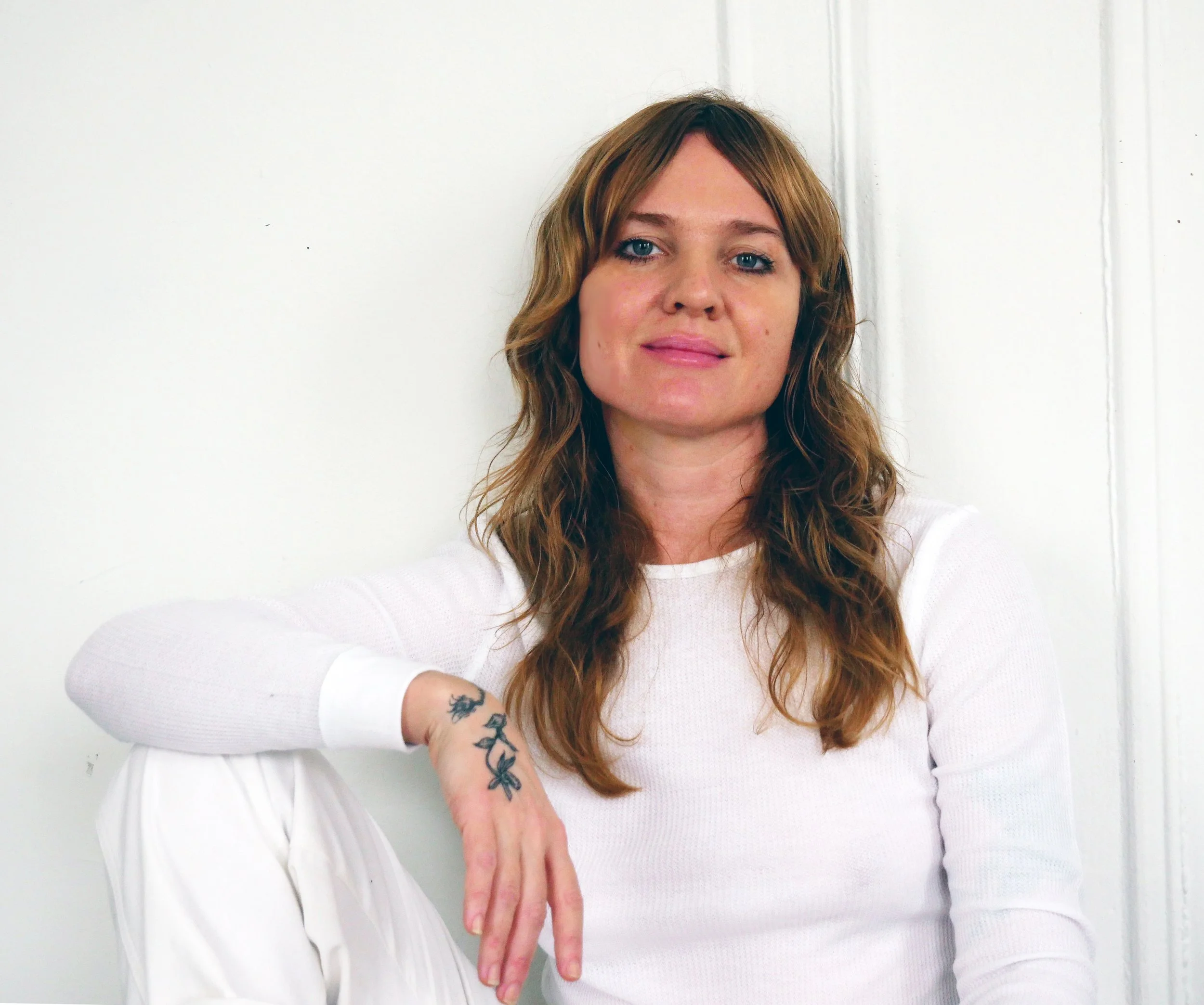 A woman with wavy reddish-brown hair sitting against a white wall, wearing a white long-sleeve top and white pants, with a tattoo on her wrist.