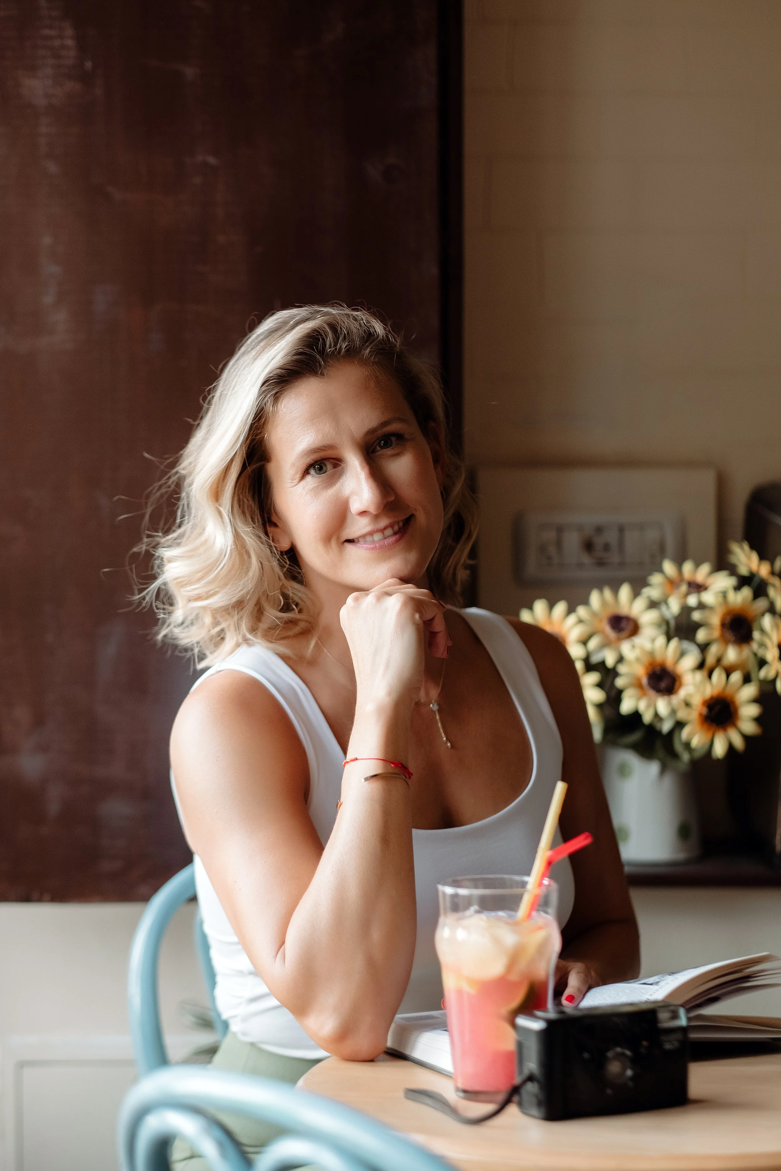 A woman sitting at a table with a book and a colorful beverage, smiling at the camera. Behind her is a potted sunflower arrangement.