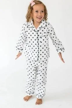 A young girl with long wavy brown hair wearing a cream-colored varsity jacket with black striped cuffs standing outdoors in a park, next to a wooden post with a gray varsity jacket hanging on it.