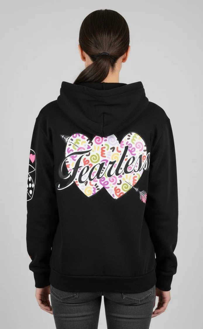 Young woman with shoulder-length dark hair wearing a pink hoodie with graphic text and illustrations, standing against a gray background.