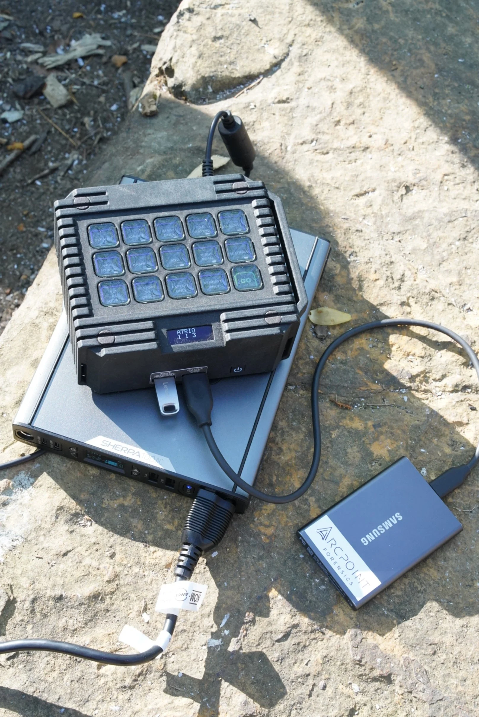 A portable electronic device with multiple small blue-tinted screens on one side, connected to a silver laptop and a Samsung external battery pack, all placed on a large rock outdoors.