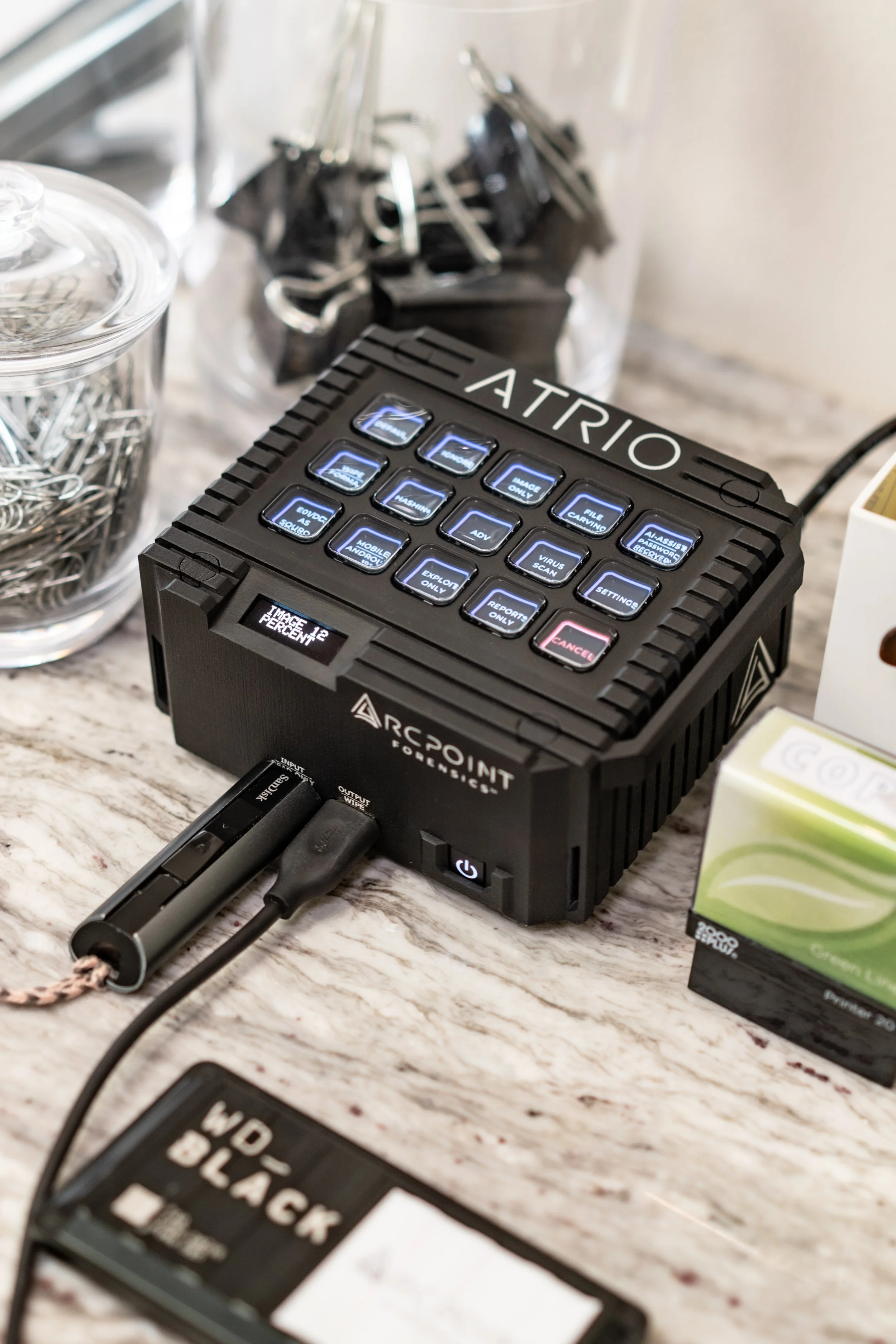 A black forensic tool labeled 'ATRIO' with illuminated buttons and a small display screen on a marble countertop, connected to a cable, with small containers of clips and a box with green packaging nearby.