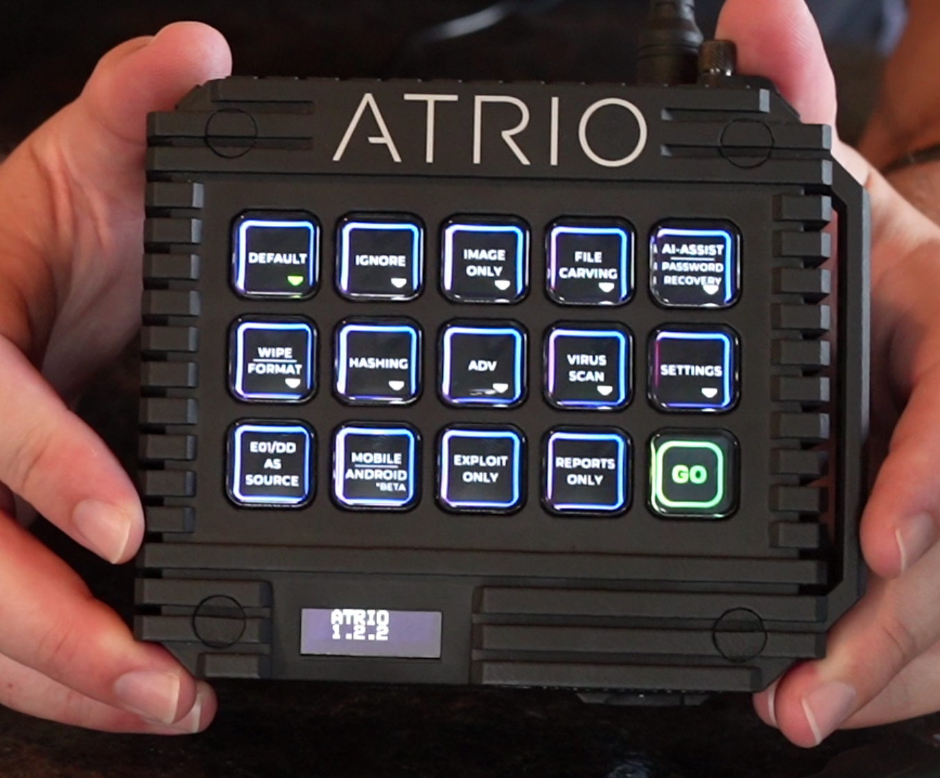 A person holding a black electronic device called ATRIO with illuminated buttons for various functions, a small digital display showing the time 1:22, and a green 'GO' button.