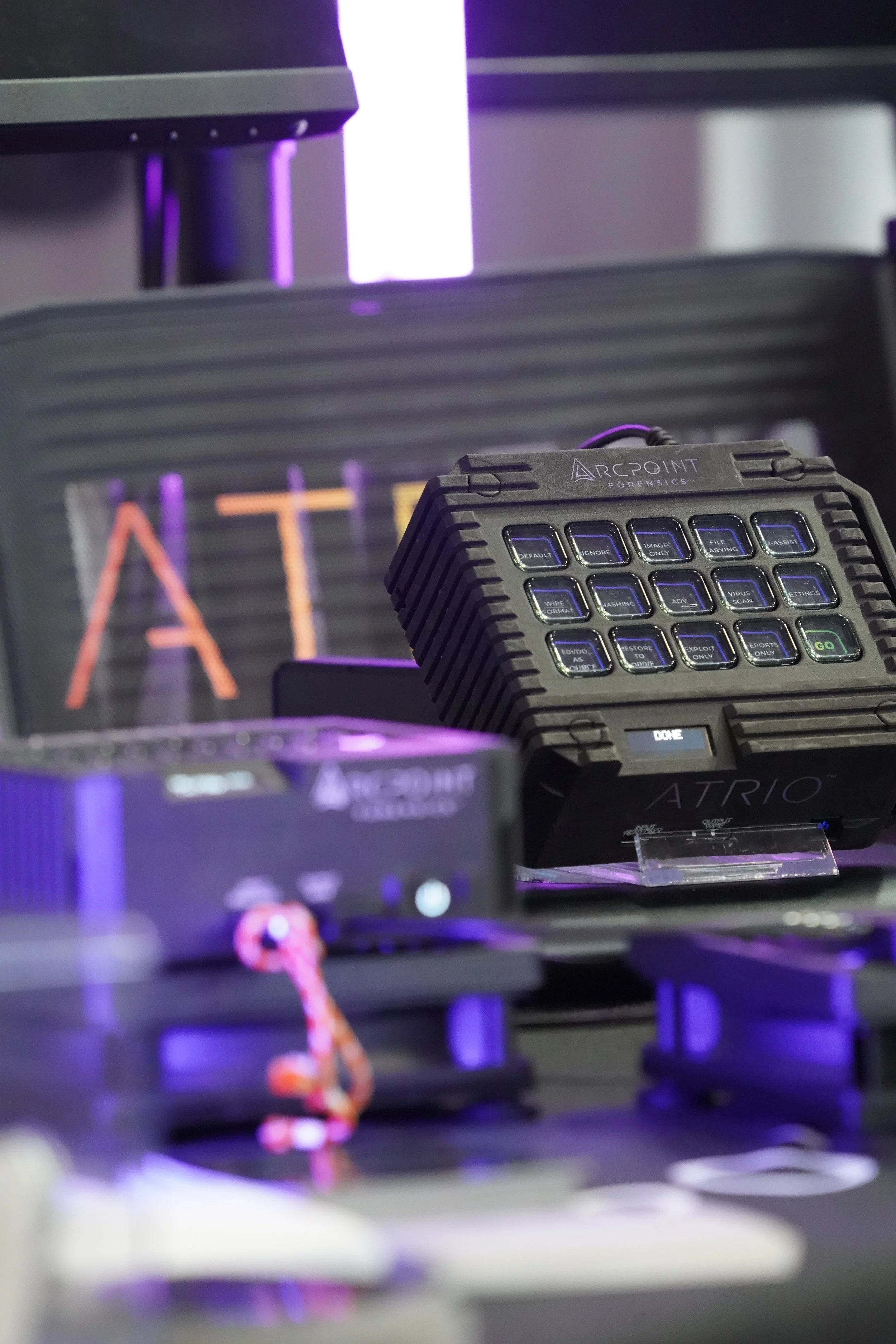 Close-up of forensic equipment on a desk, including a handheld device with multiple buttons, a small monitor with illuminated text 'ATT' in red, and blurred objects with purple lighting in the background.