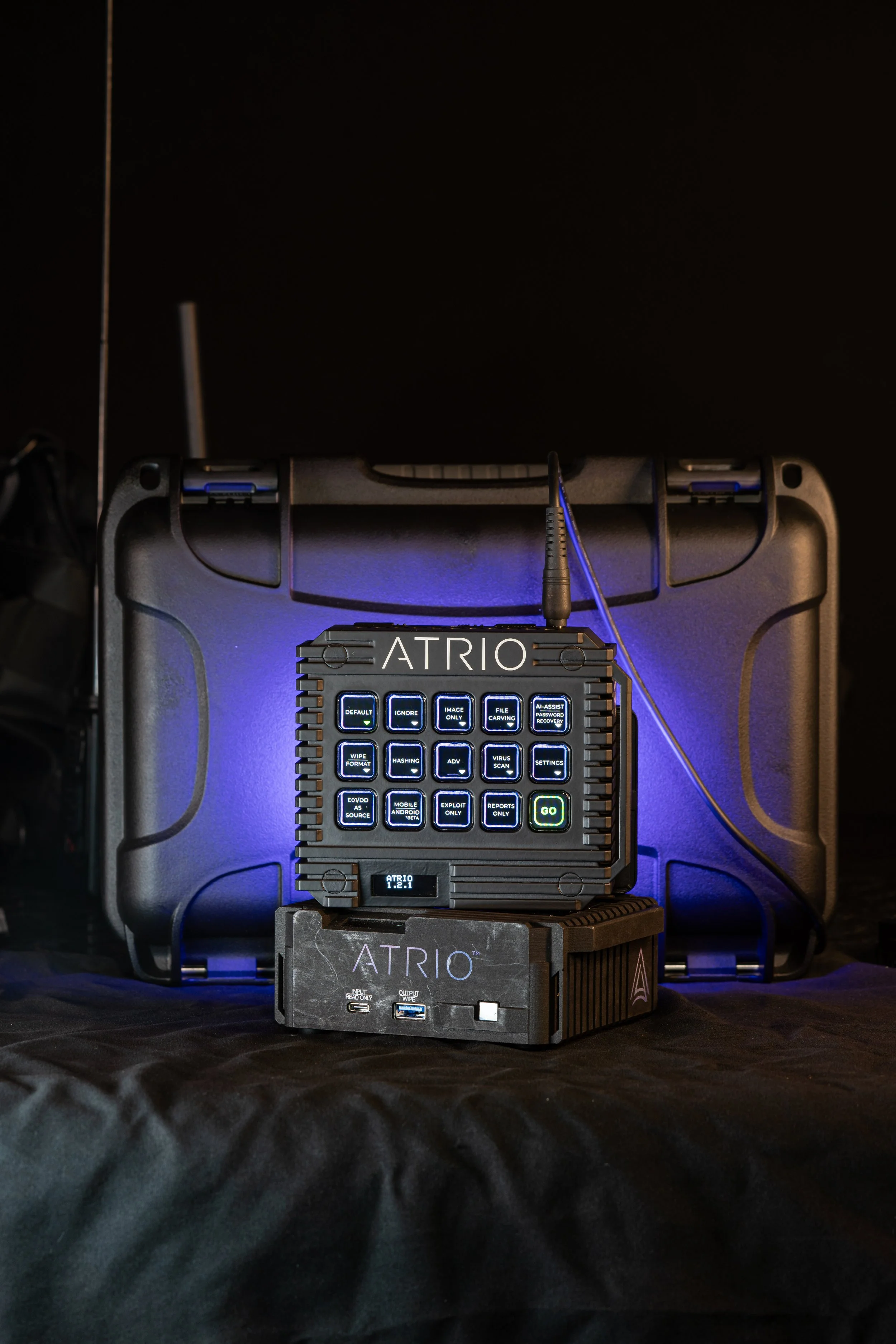 A portable electronic device labeled 'ATRIO' with illuminated buttons, placed on a black surface with a black case and wire in the background.