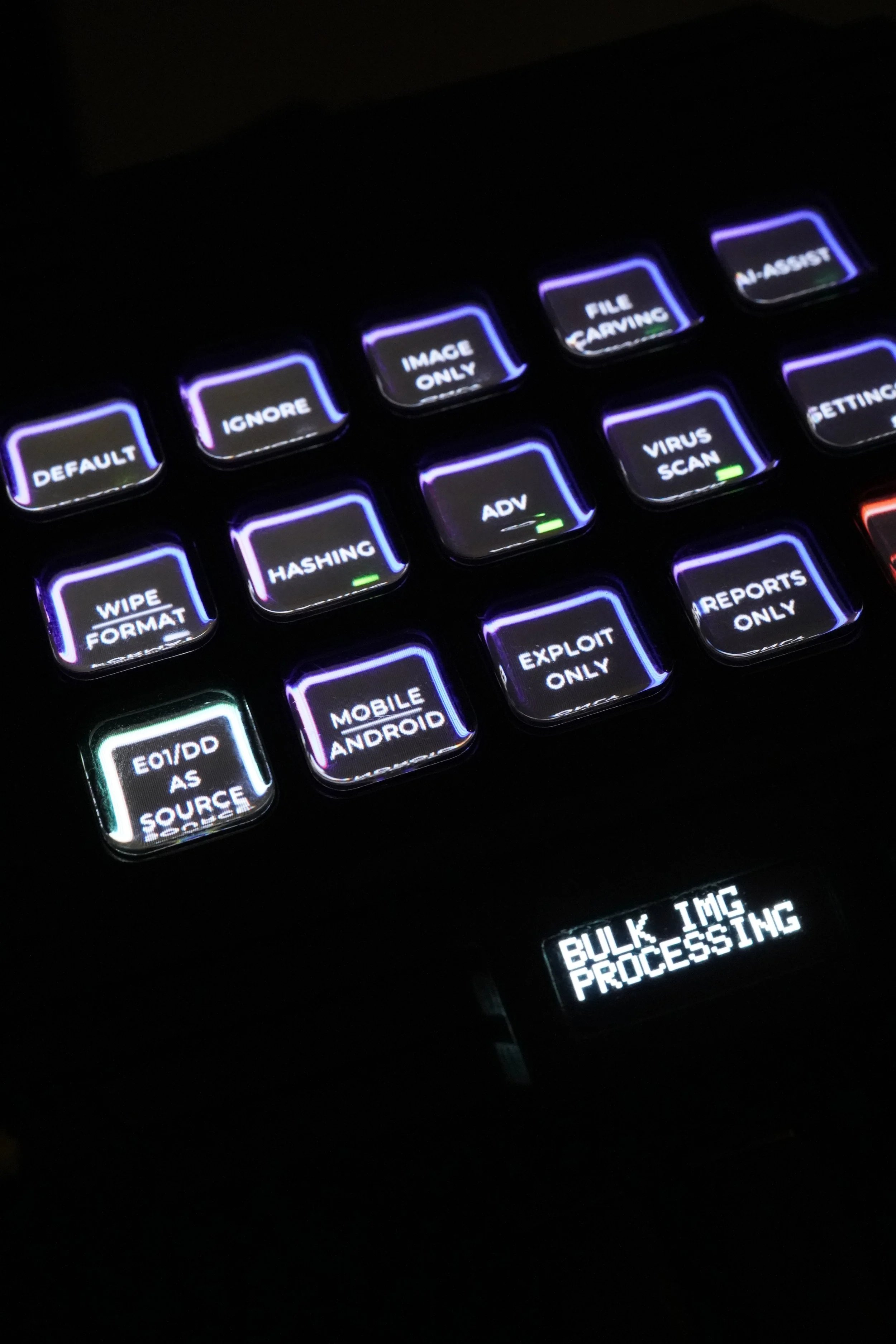 A close-up photograph of a digital keypad with illuminated keys, including labels such as DEFAULT, IGNORE, IMAGE ONLY, FILE CARVING, AI-ASSIST, WIPE FORMAT, HASHING, ADV, VIRUS SCAN, SETTINGS, E01/DD AS SOURCE, MOBILE ANDROID, EXPLOIT ONLY, REPORTS ONLY, and a small screen displaying "BULK IMG PROCESSING."