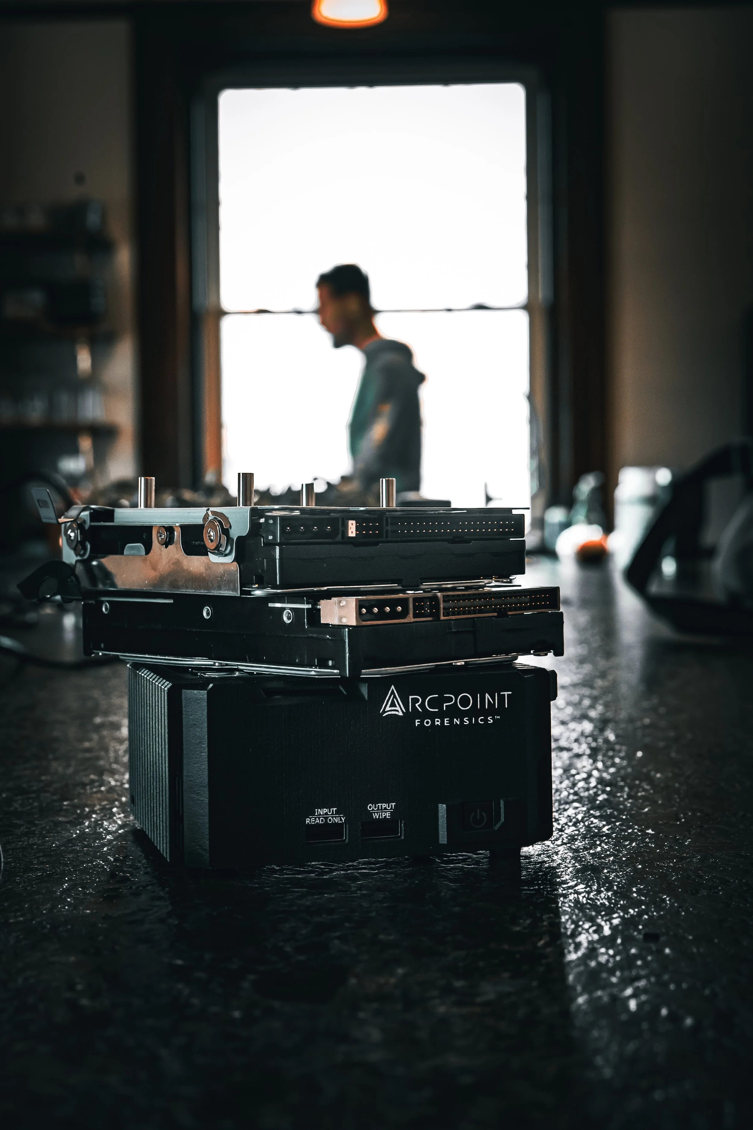 Close-up of forensic hard drives and equipment labeled ArcPoint Forensics on a dark table in a dimly lit room, with a man working at a desk blurred in the background near a window.