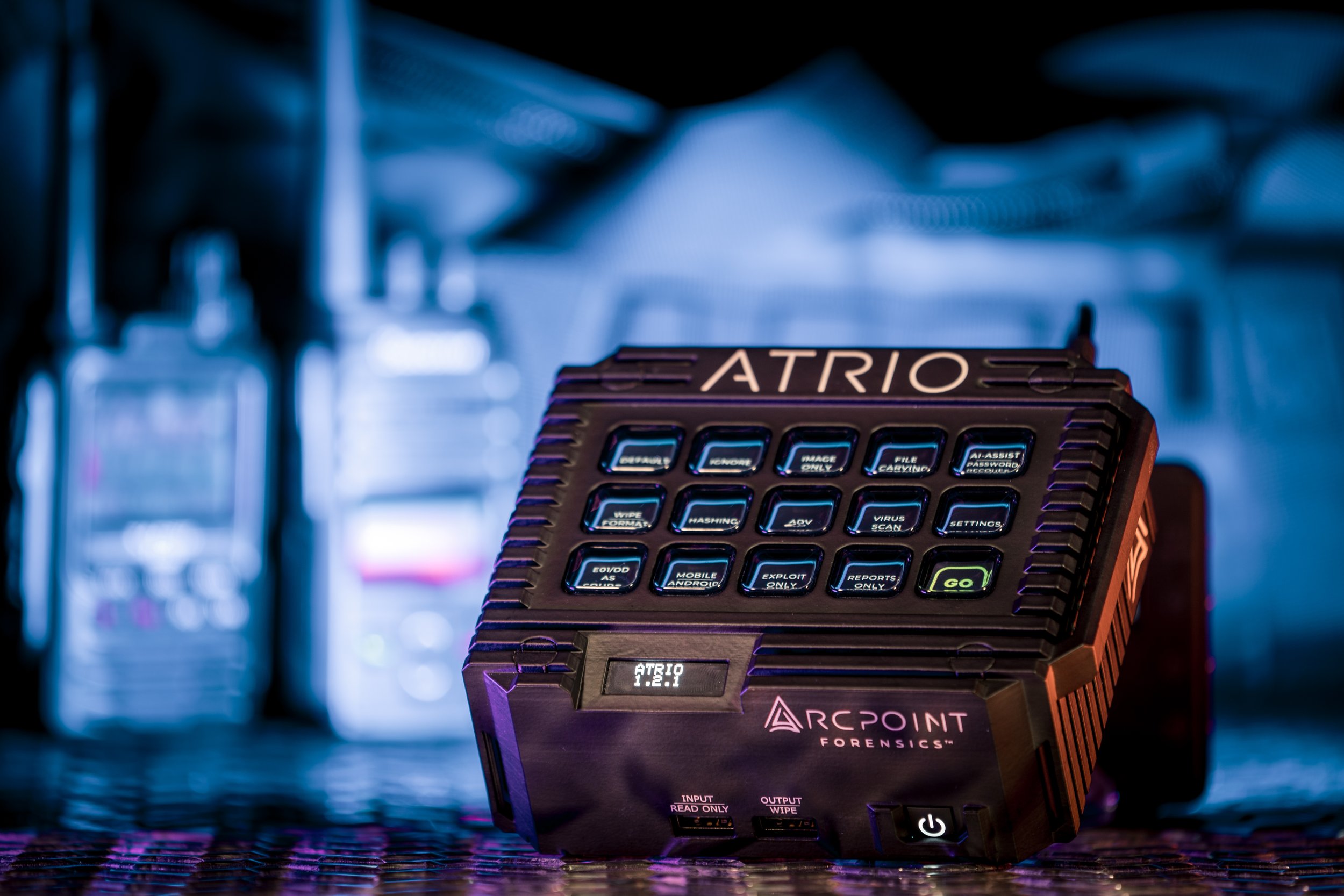 A dark, portable forensic device labeled 'ATRIO' by Arcpoint Forensics, with a colorful backlit keypad and a small screen displaying 'ATRIO 1.2.1,' sitting on a metallic surface with blurred laboratory equipment in the background.