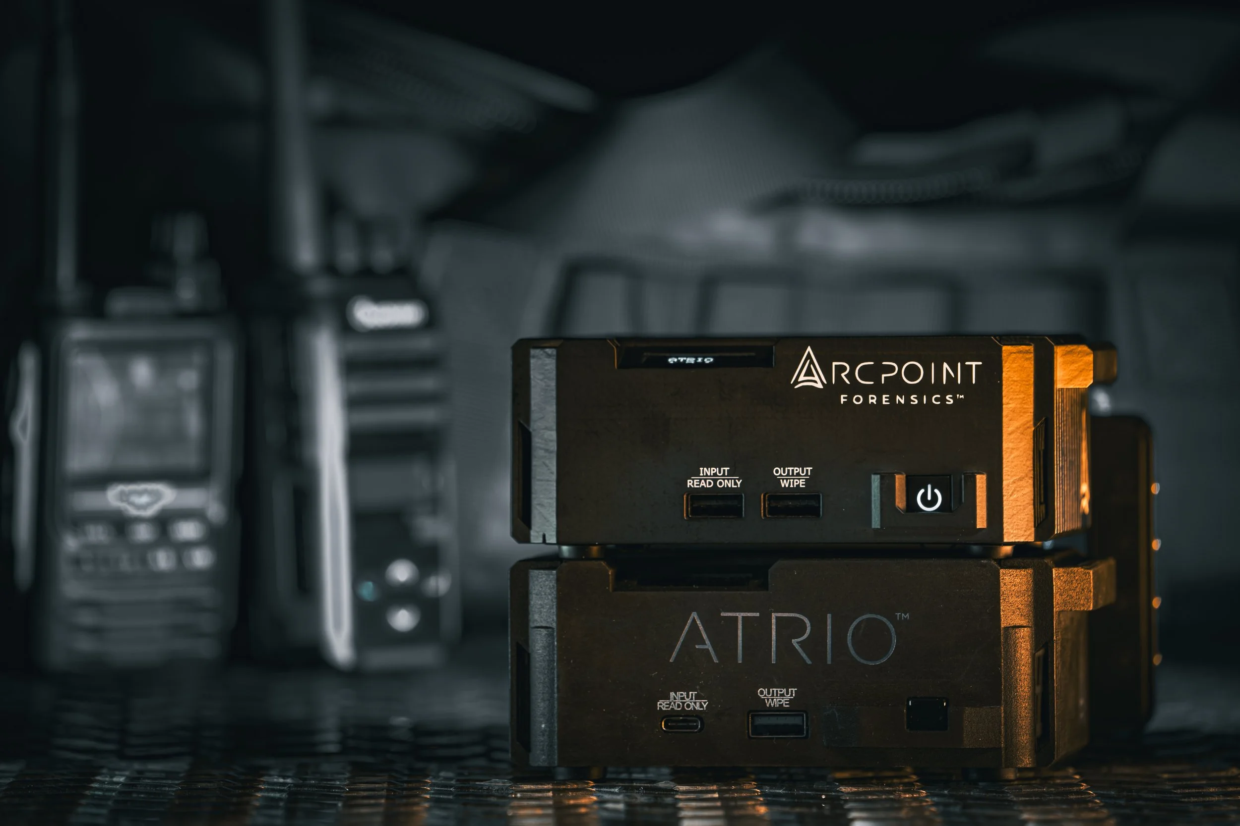 Close-up of forensic equipment labeled 'Arcpoint Forensic' and 'ATRIO' with various ports, lights, and buttons, placed on a dark textured surface, with blurred tools in the background.