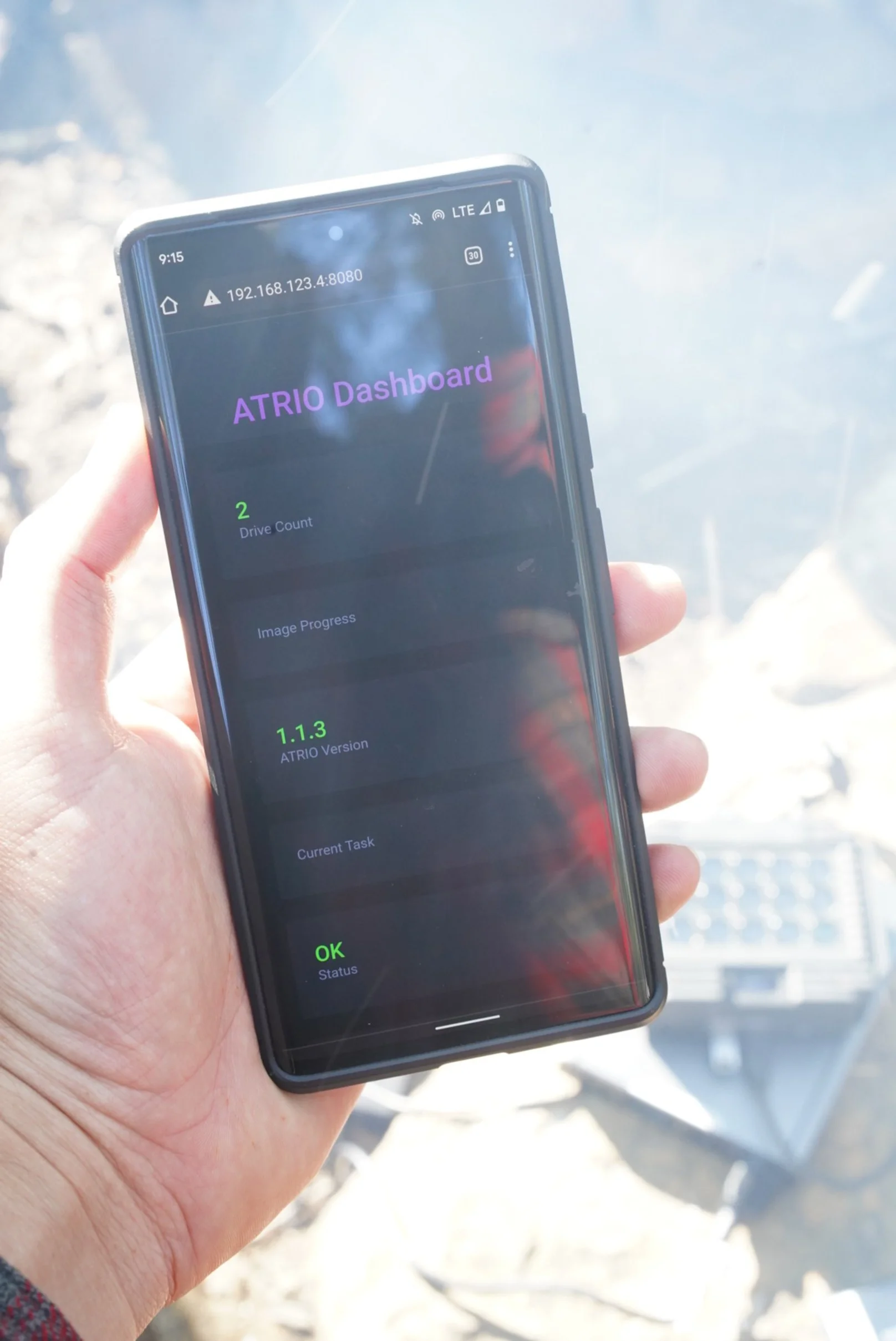 A person holding a smartphone displaying an ATRIO Dashboard with drive count of 2, version 1.1.3, and status 'OK'.