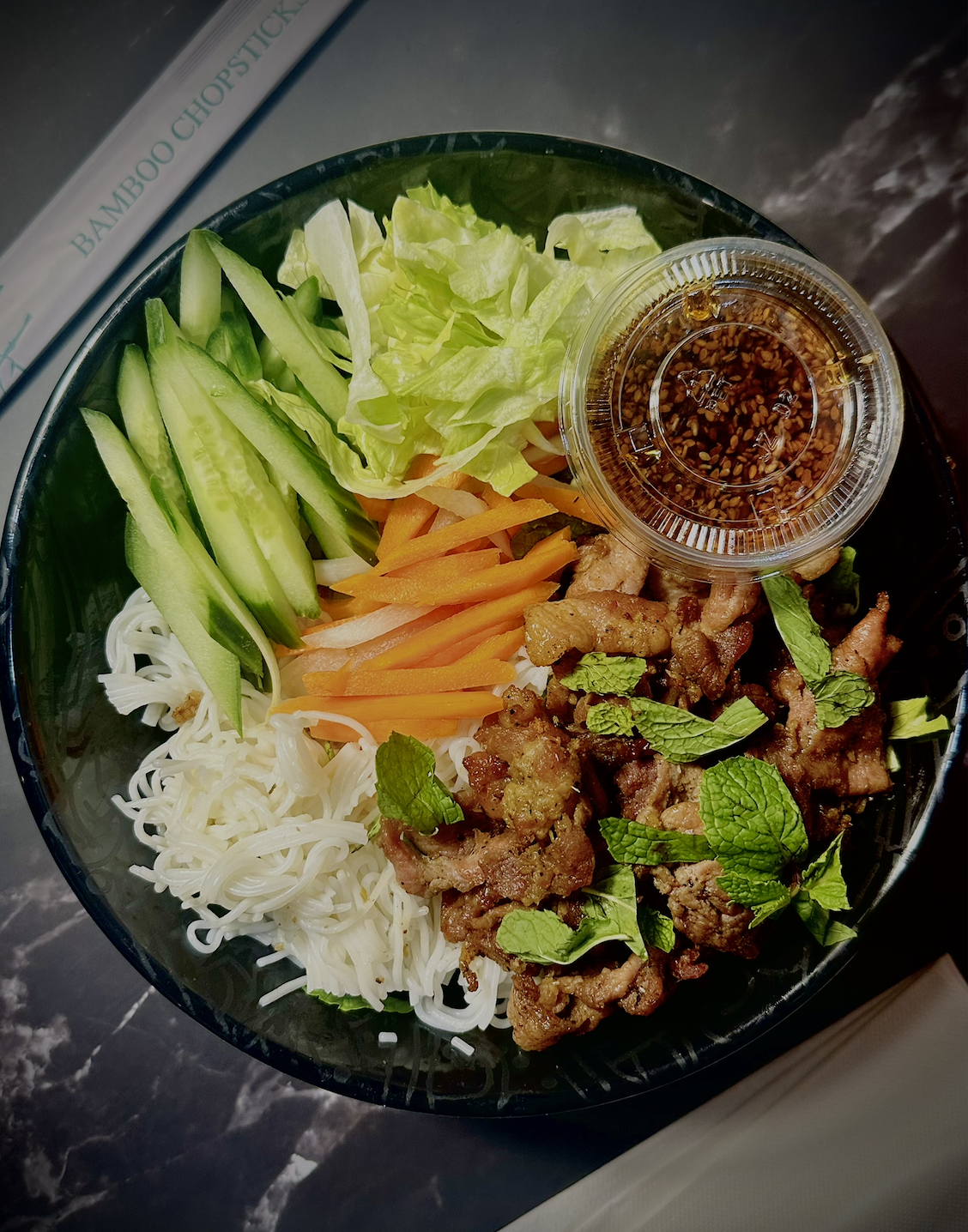 Bowl of Asian noodle salad with sliced cucumbers, lettuce, shredded carrots, vermicelli noodles, grilled meat, fresh mint, and a small cup of dipping sauce.