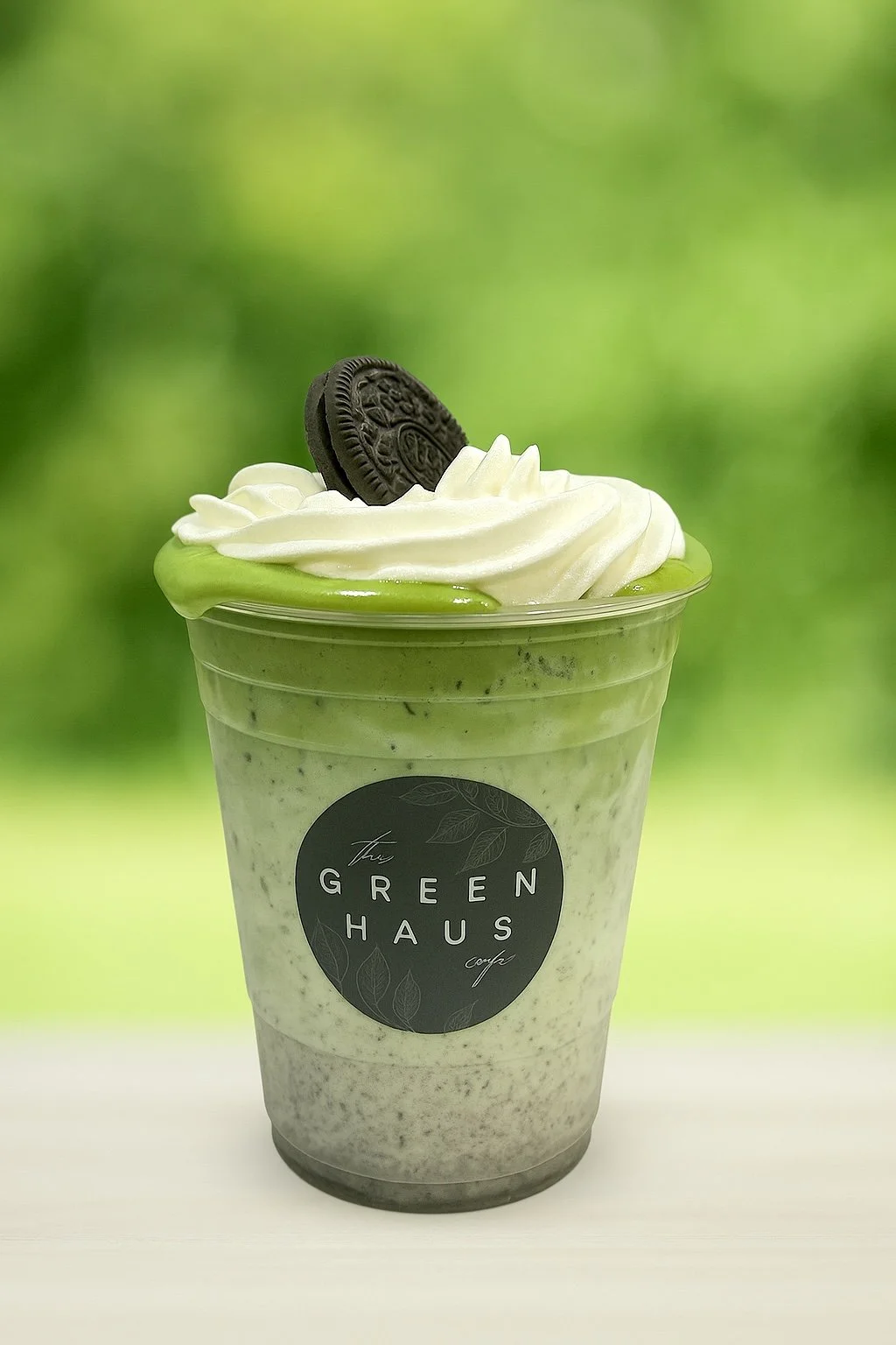 Green matcha smoothie in a clear plastic cup with whipped cream and an Oreo cookie on top, against a blurred green background.