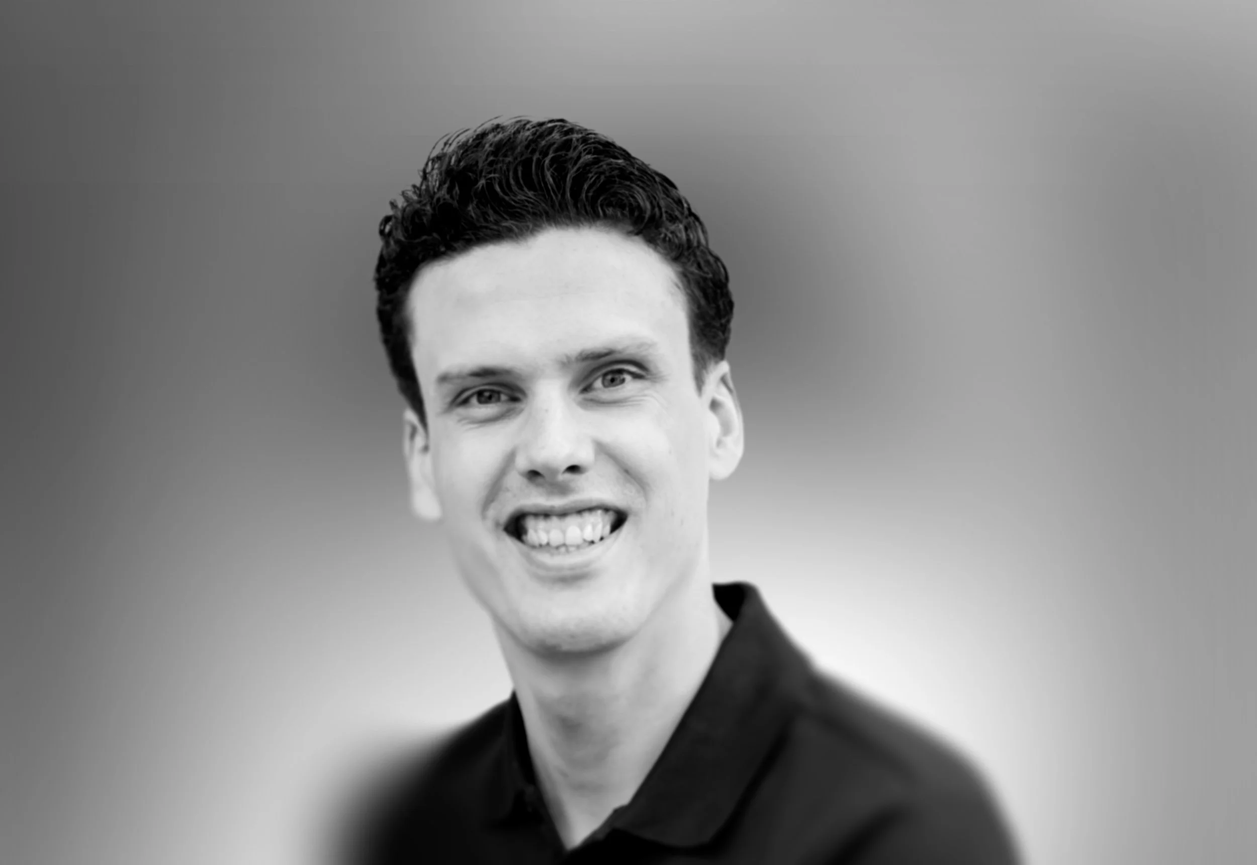 Een zwart-wit foto van een lachende man met donker, gekamd haar, gekleed in een donker pak.