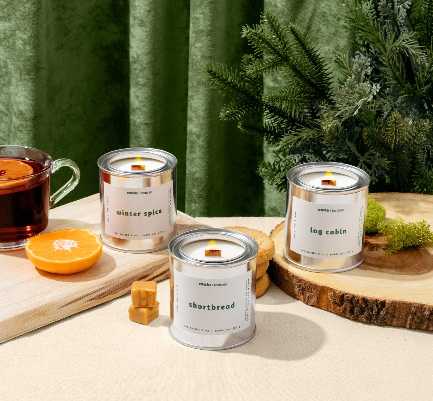 Three scented candles with labels reading 'winter spice,' 'log cabin,' and 'shortbread' are arranged on a table with a jar of tea, a half orange, some cookies, and greenery in the background.
